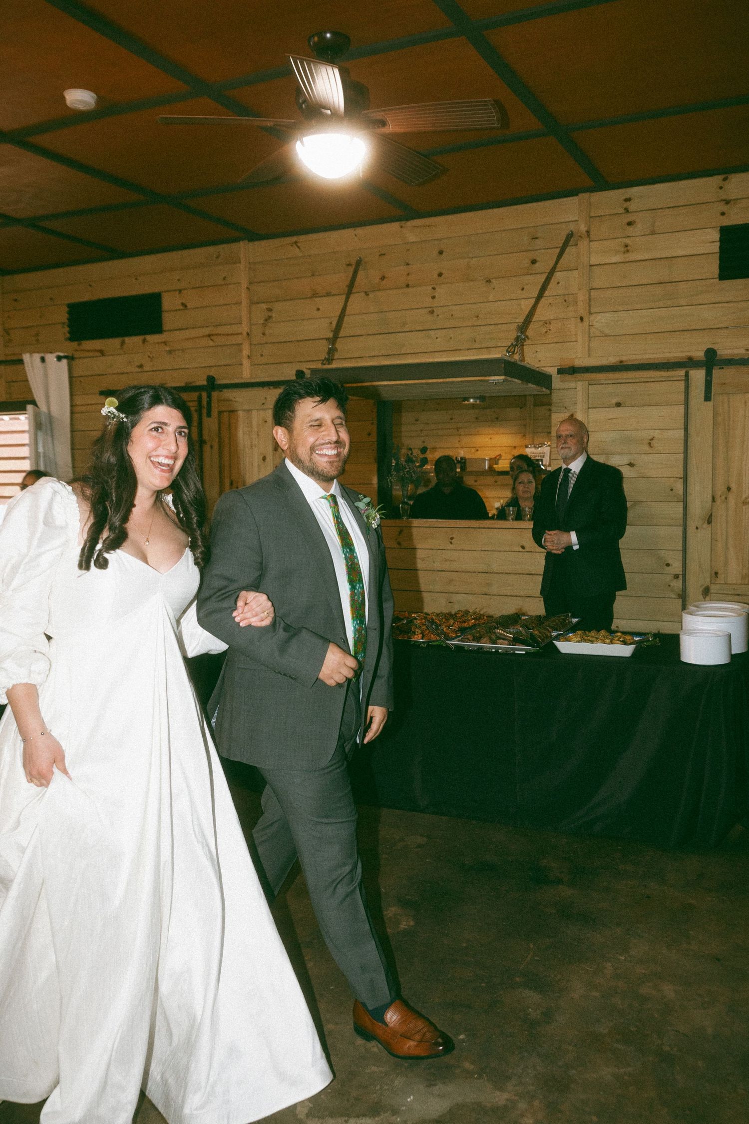 A joyful bride in white gown and groom in gray suit with green tie make their grand entrance at a rustic barn wedding reception.