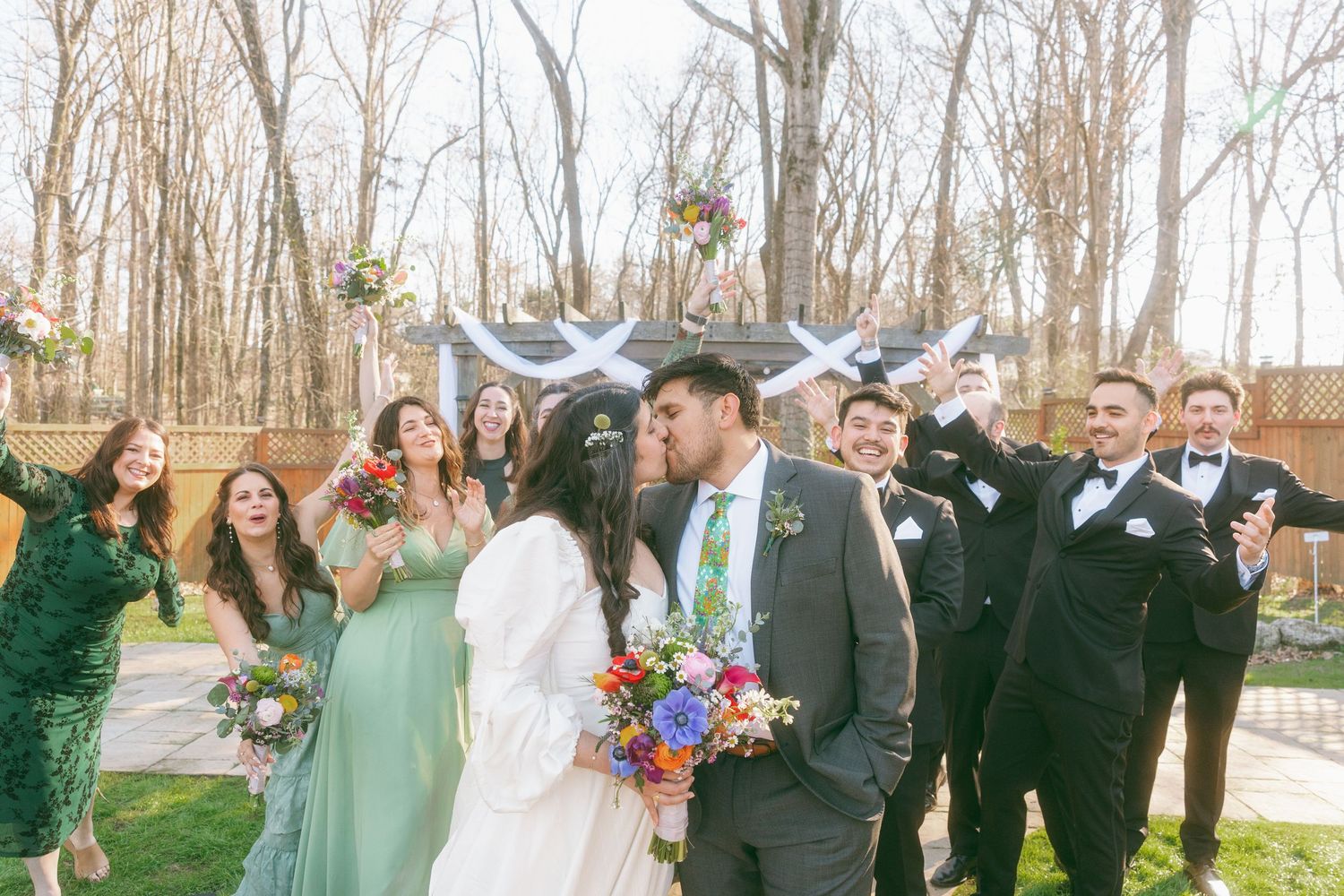 Bride and groom share a kiss surrounded by smiling wedding party in colorful attire at an outdoor ceremony.