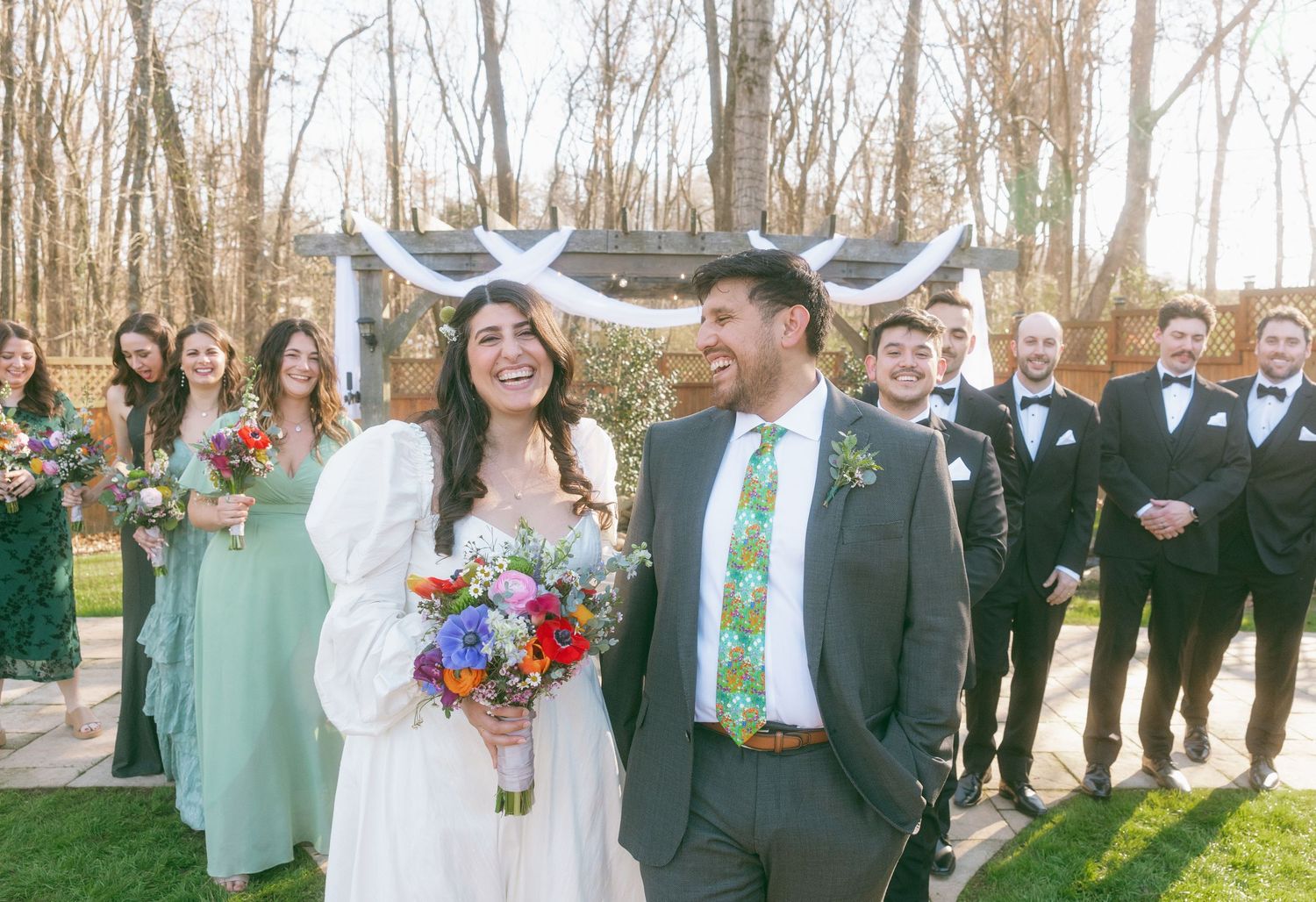 Smiling bride and groom stand with wedding party in sage green and black tuxedos at outdoor ceremony.