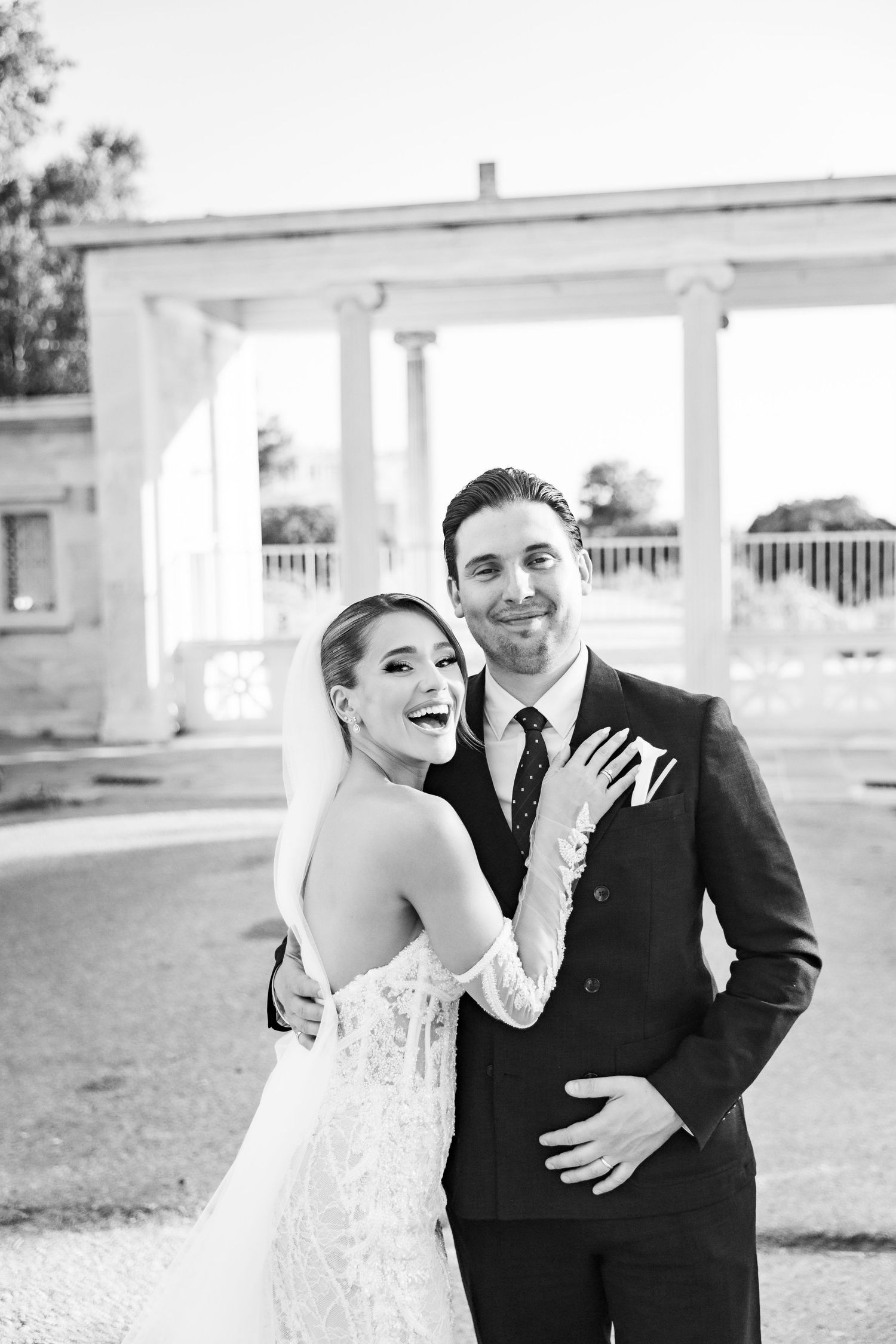 Black and white photo of a joyful bride and groom posing together outdoors near elegant columns on their wedding day.
