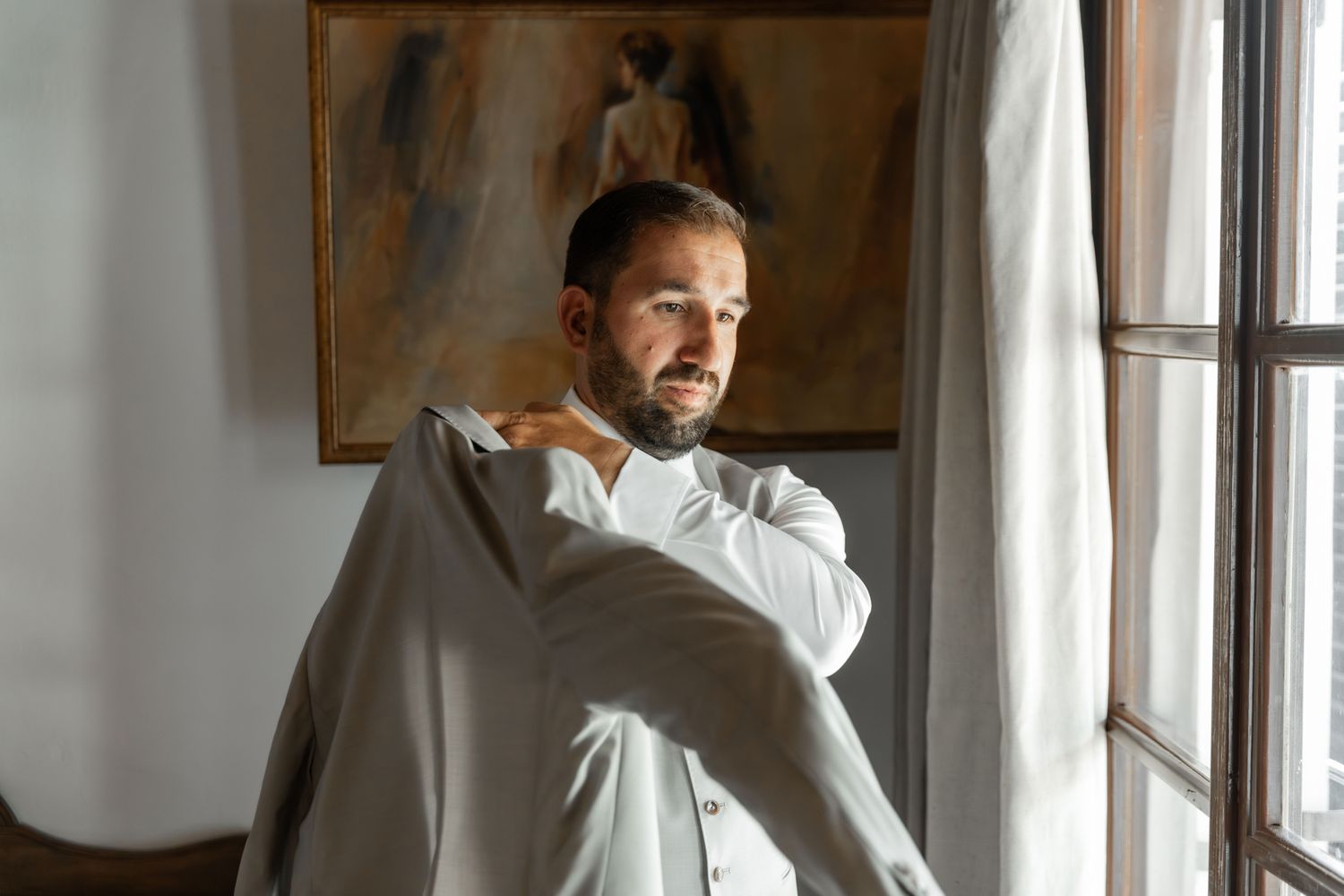 A person in formal attire stands near a window in a minimalist interior setting.