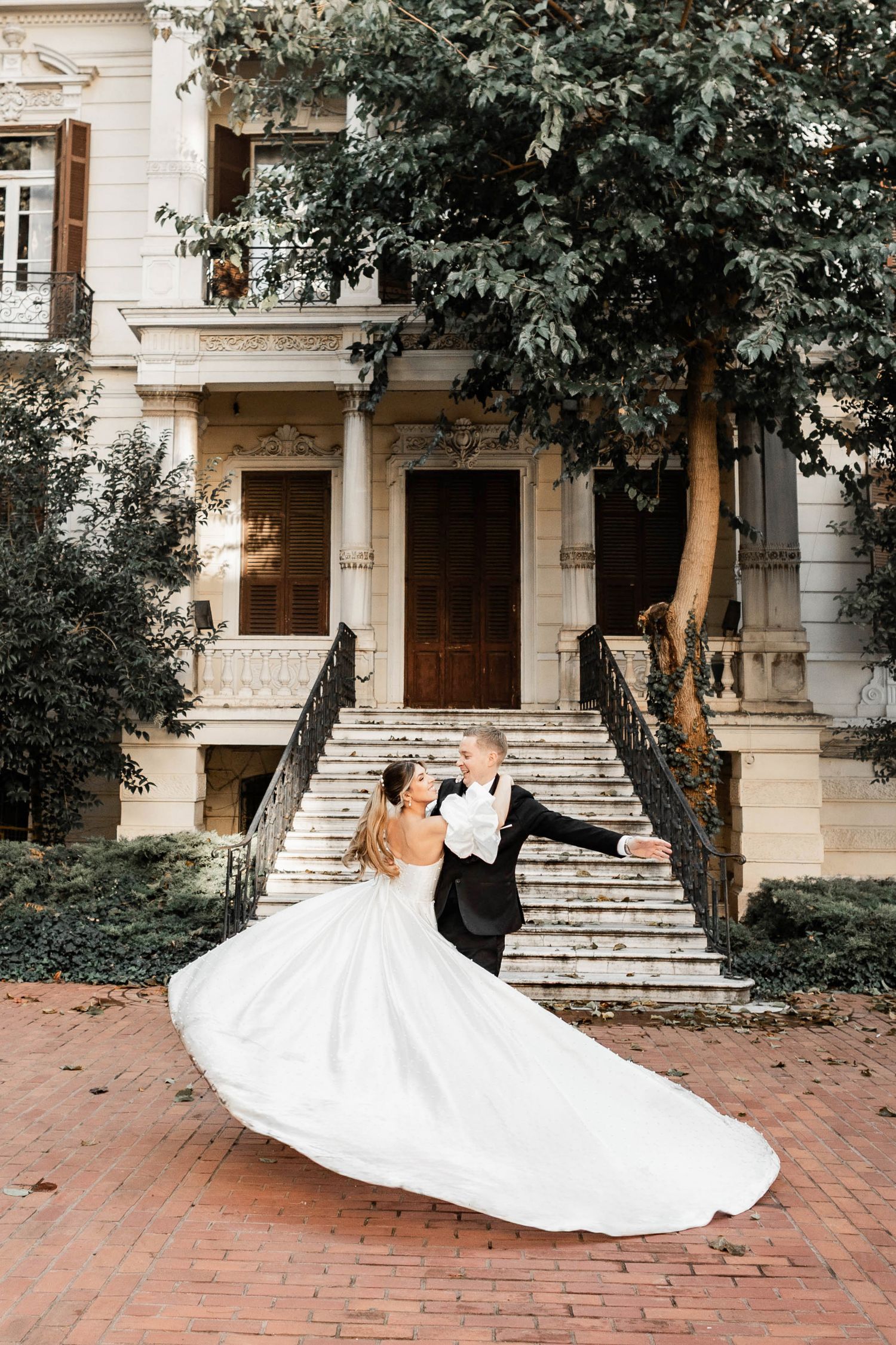 Romantic dip pose of newlyweds on brick pathway with elegant mansion backdrop.
