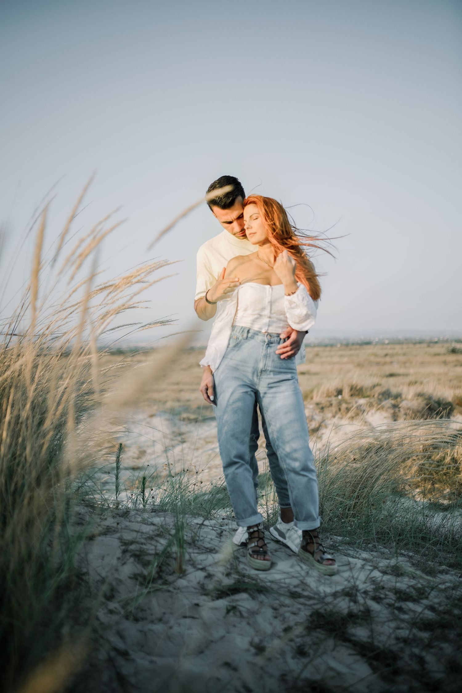 A couple embraces on a sandy beach with tall grass swaying in the golden sunset light.