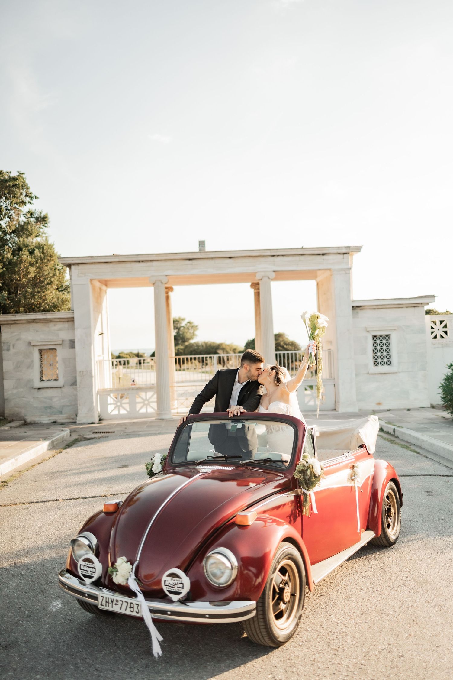 Newlywed couple kissing in a red vintage VW Beetle convertible decorated with white ribbons outside elegant venue.