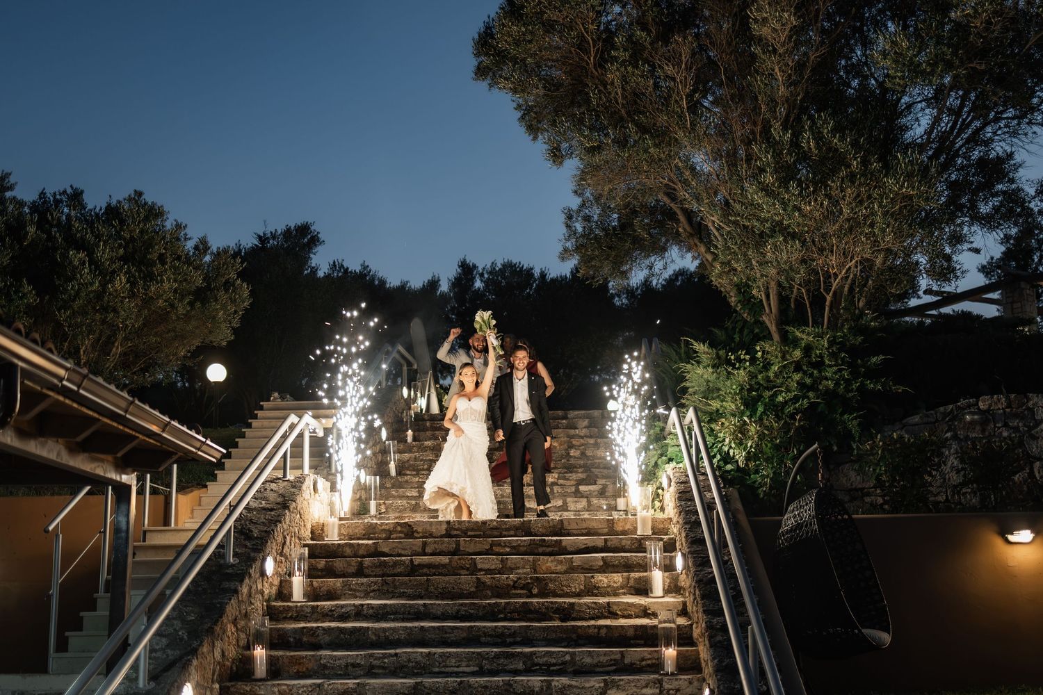 Bride and groom descend sparkler-lined stone steps at night, celebrating their wedding exit under a dark blue sky.