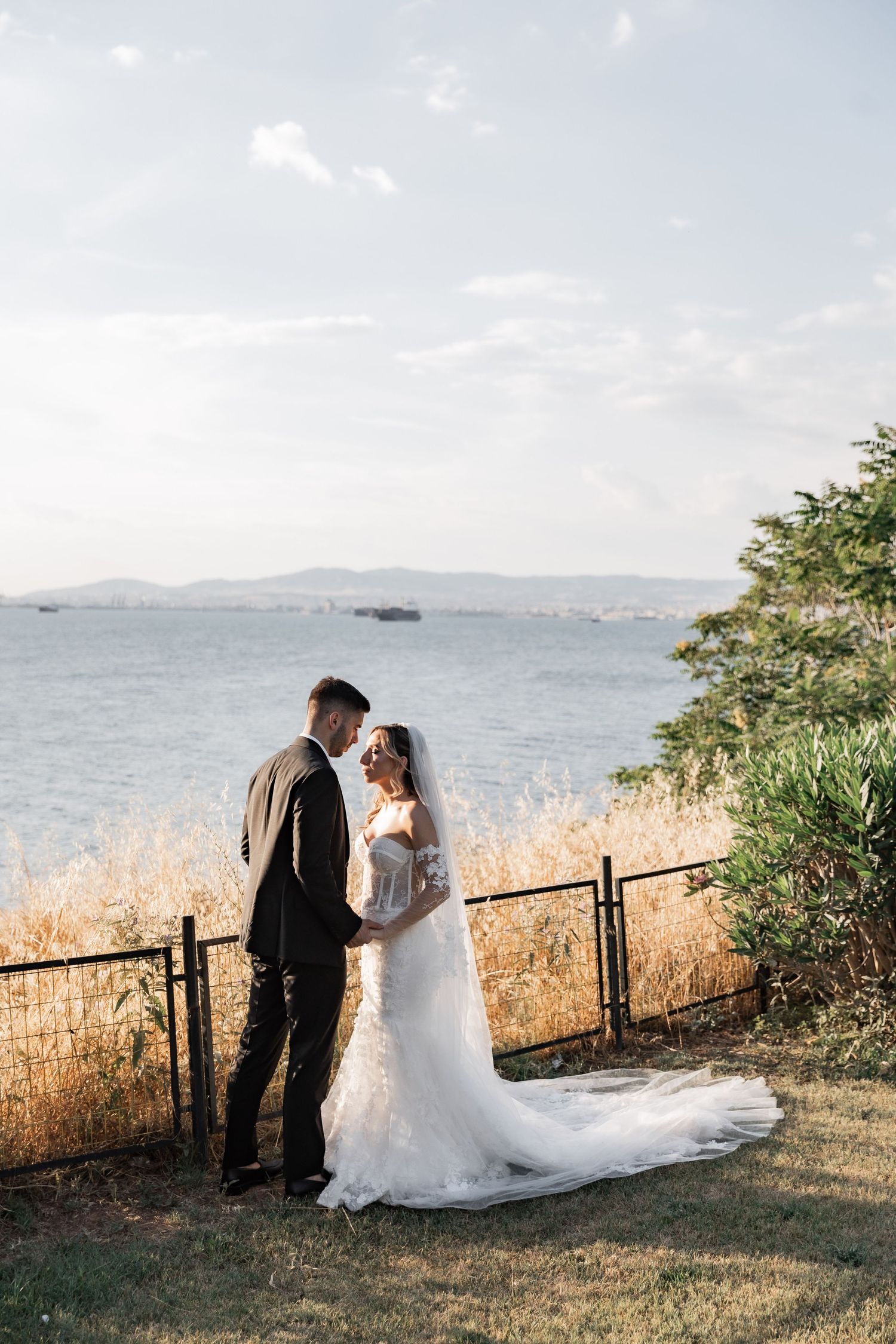 Bride and groom holding hands by the sea, wearing wedding dress and suit, with mountains in the background.
