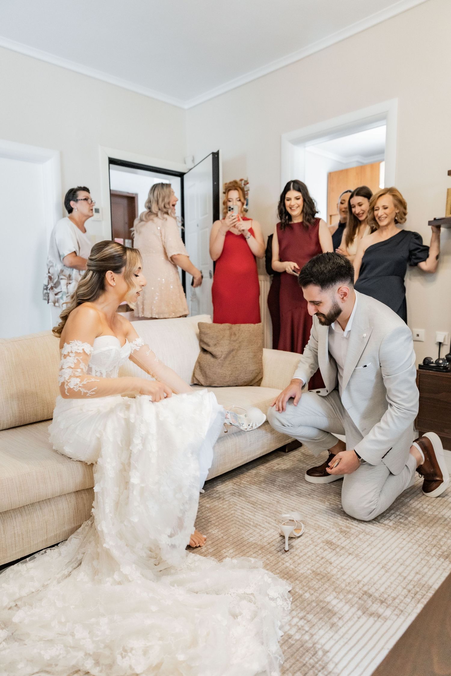 Groom kneeling before bride on sofa, surrounded by smiling wedding guests in elegant attire during bridal preparation.