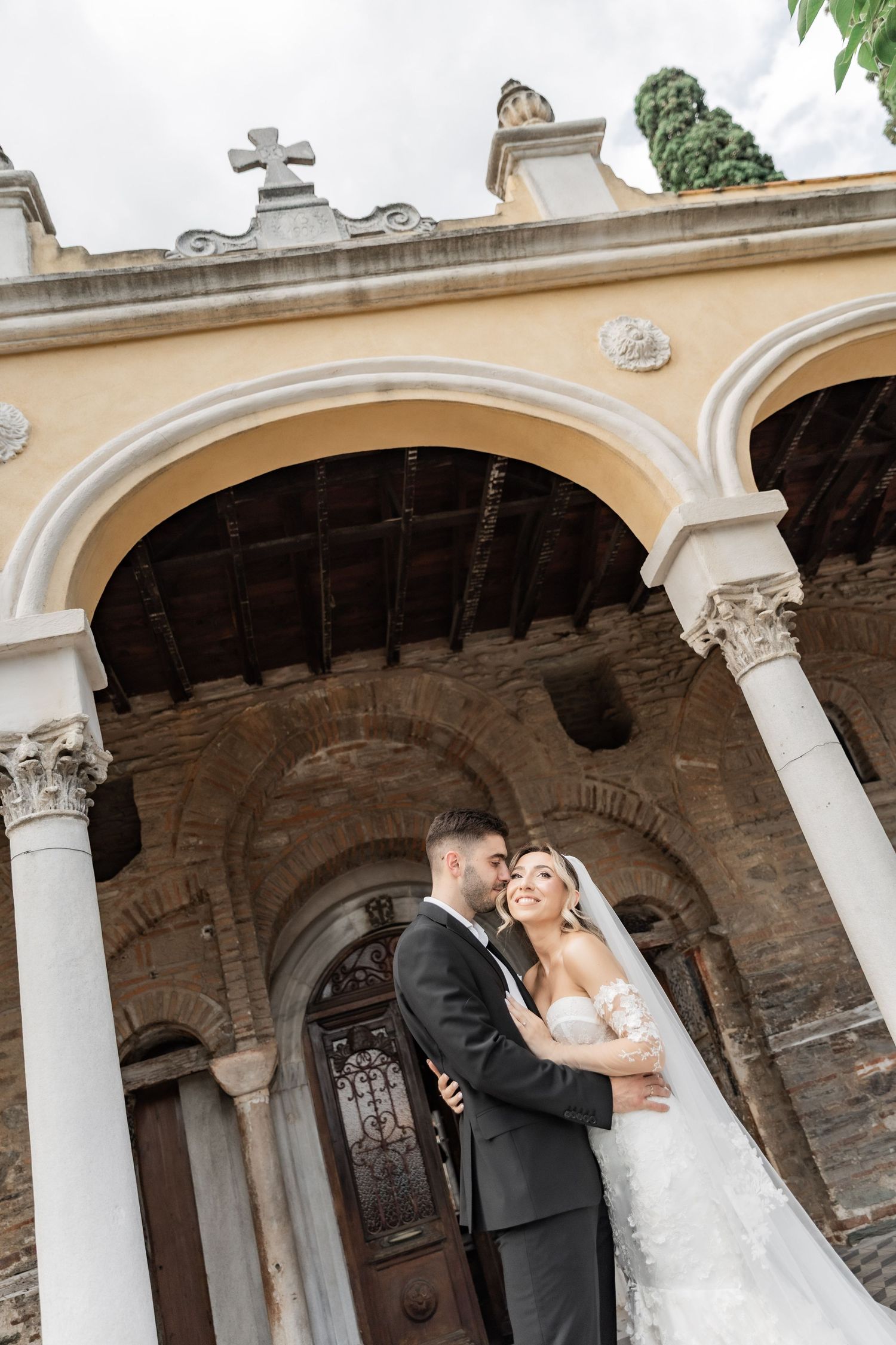 Bride and groom embrace before a historic church with white arched columns and a cross, capturing a romantic wedding moment.