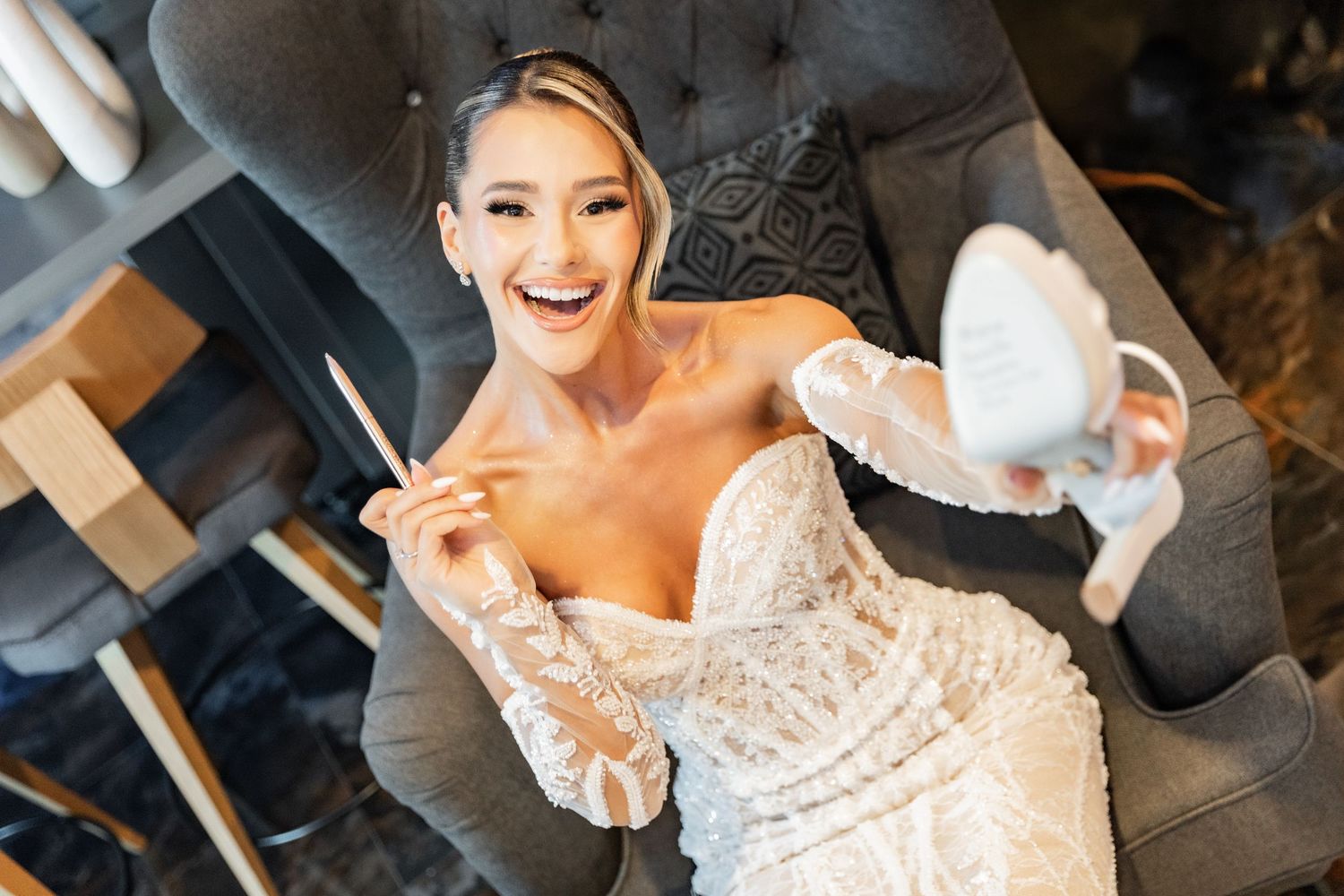 Bride in lace wedding gown laughing while holding white heels, seated in gray chair during bridal prep.