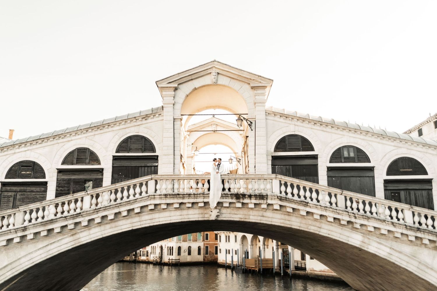 Iconic Rialto Bridge in Venice with its grand white stone arches and ornate architectural details against a bright sky.