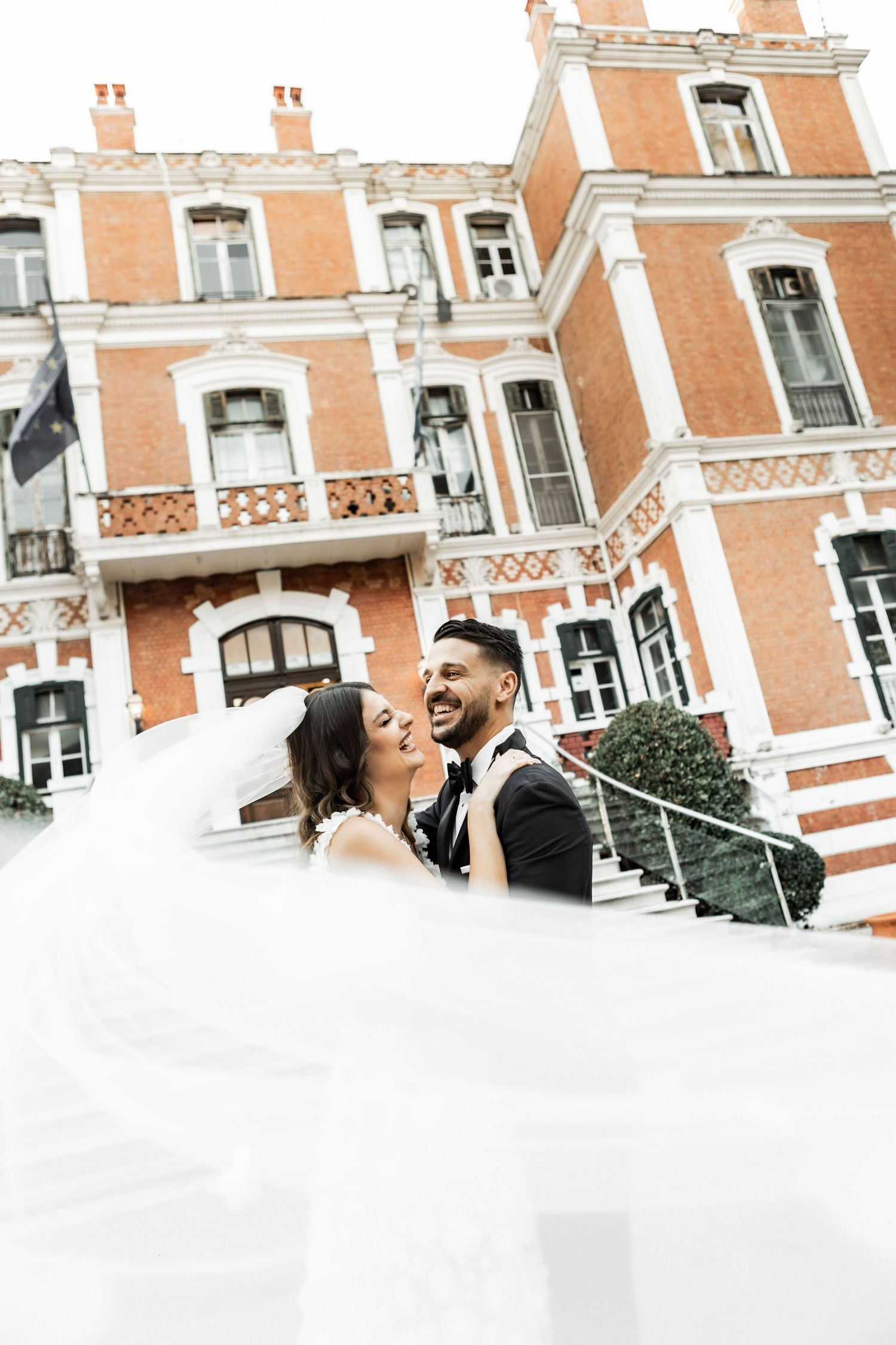 A romantic wedding portrait in front of a grand orange brick building with elegant architectural details.