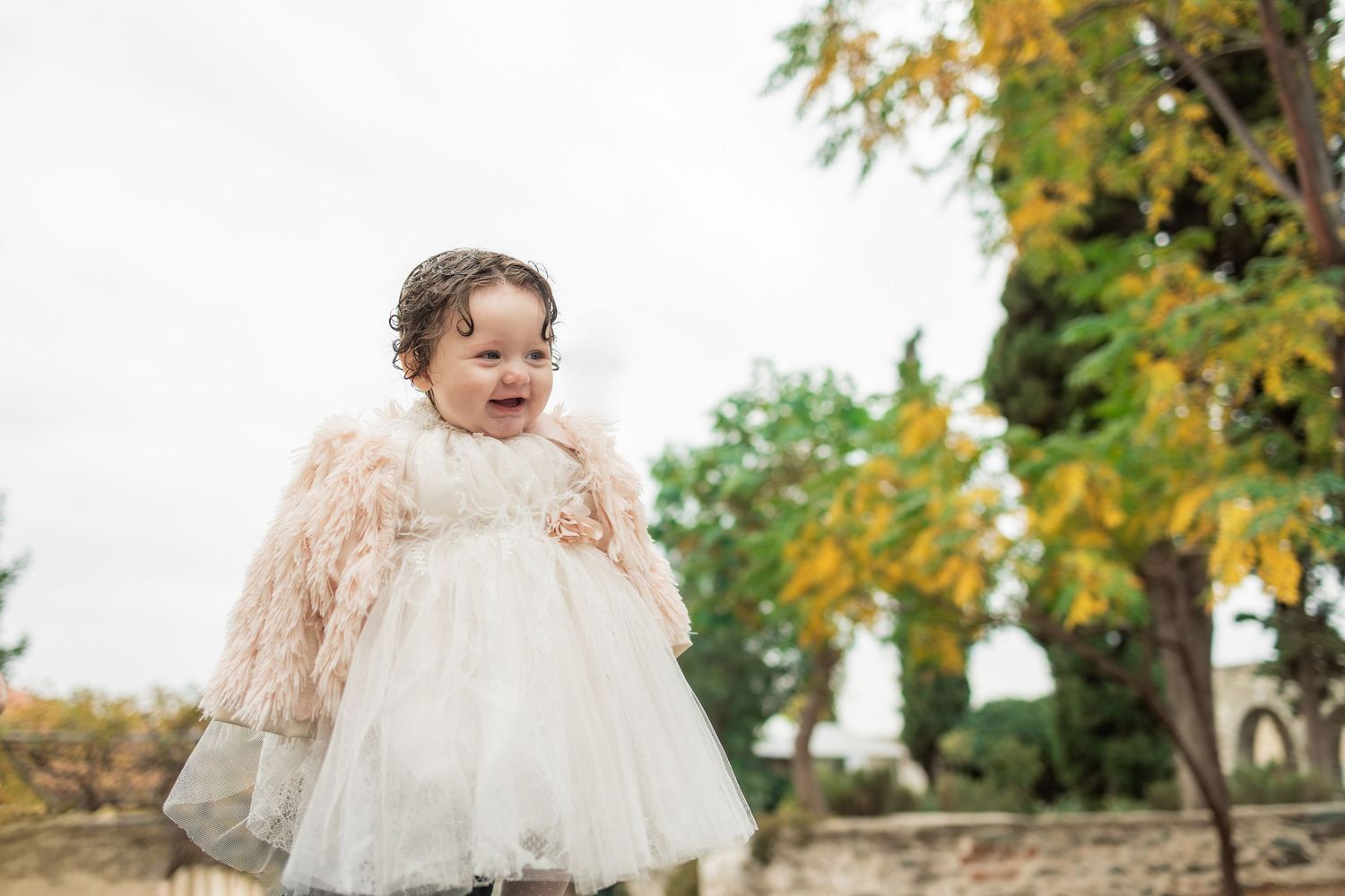 A baby in a white lace dress laughs while sitting outdoors among autumn trees and falling leaves.