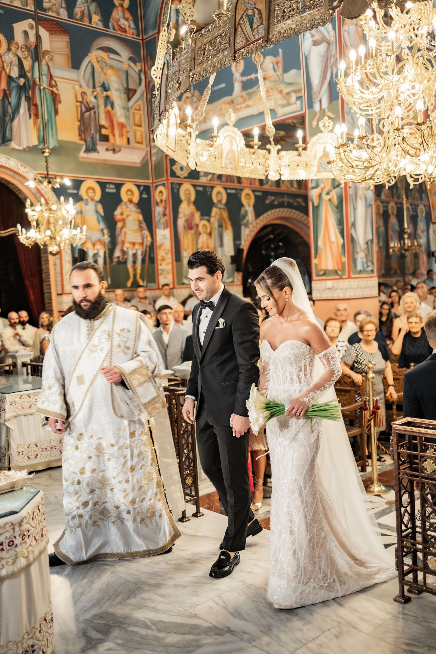 Bride and groom walk down the aisle in an ornate Orthodox church with golden chandeliers and colorful frescoes.