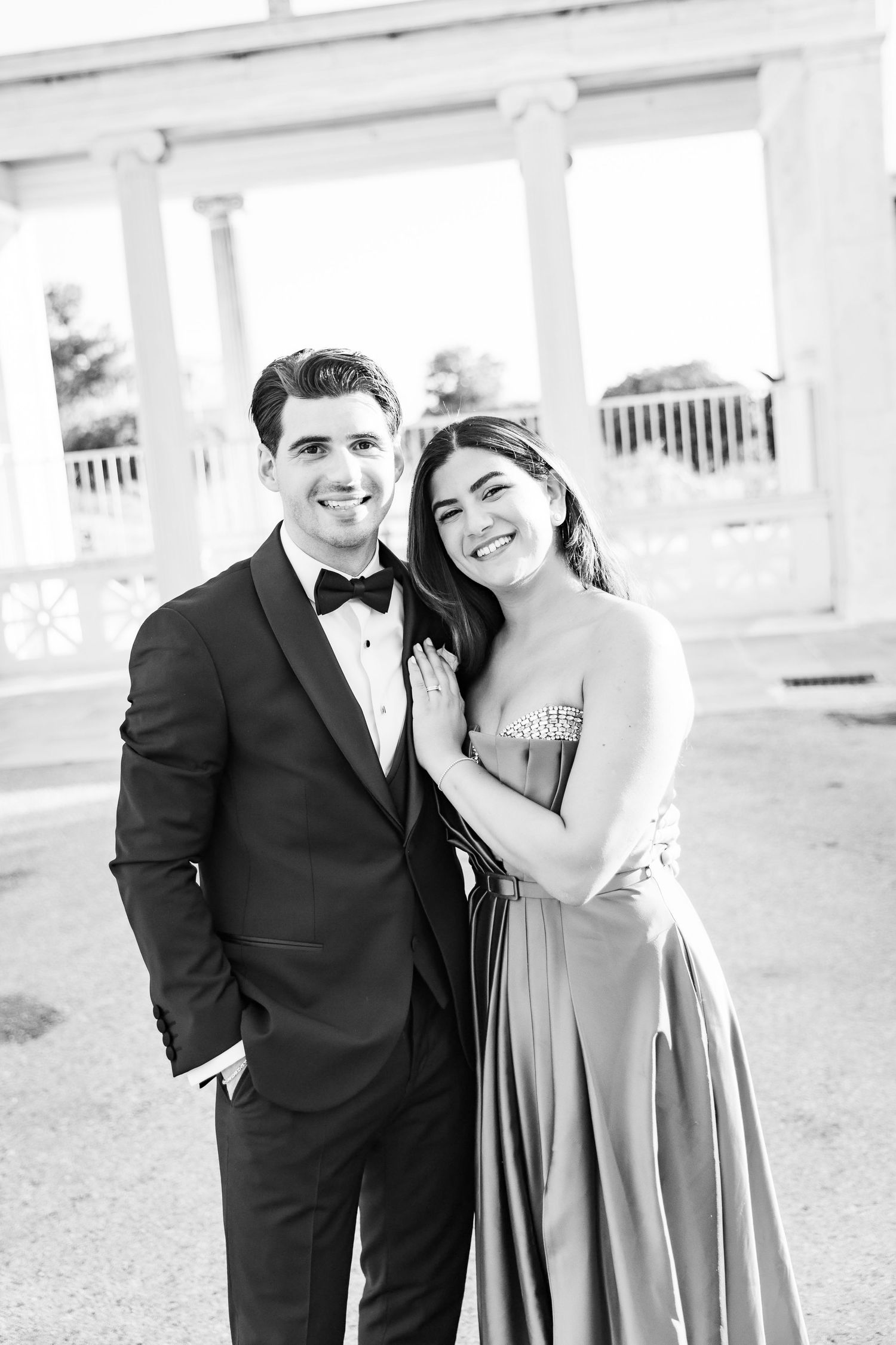Elegant couple posing in black and white, man in tuxedo and woman in strapless gown, smiling at a formal venue.