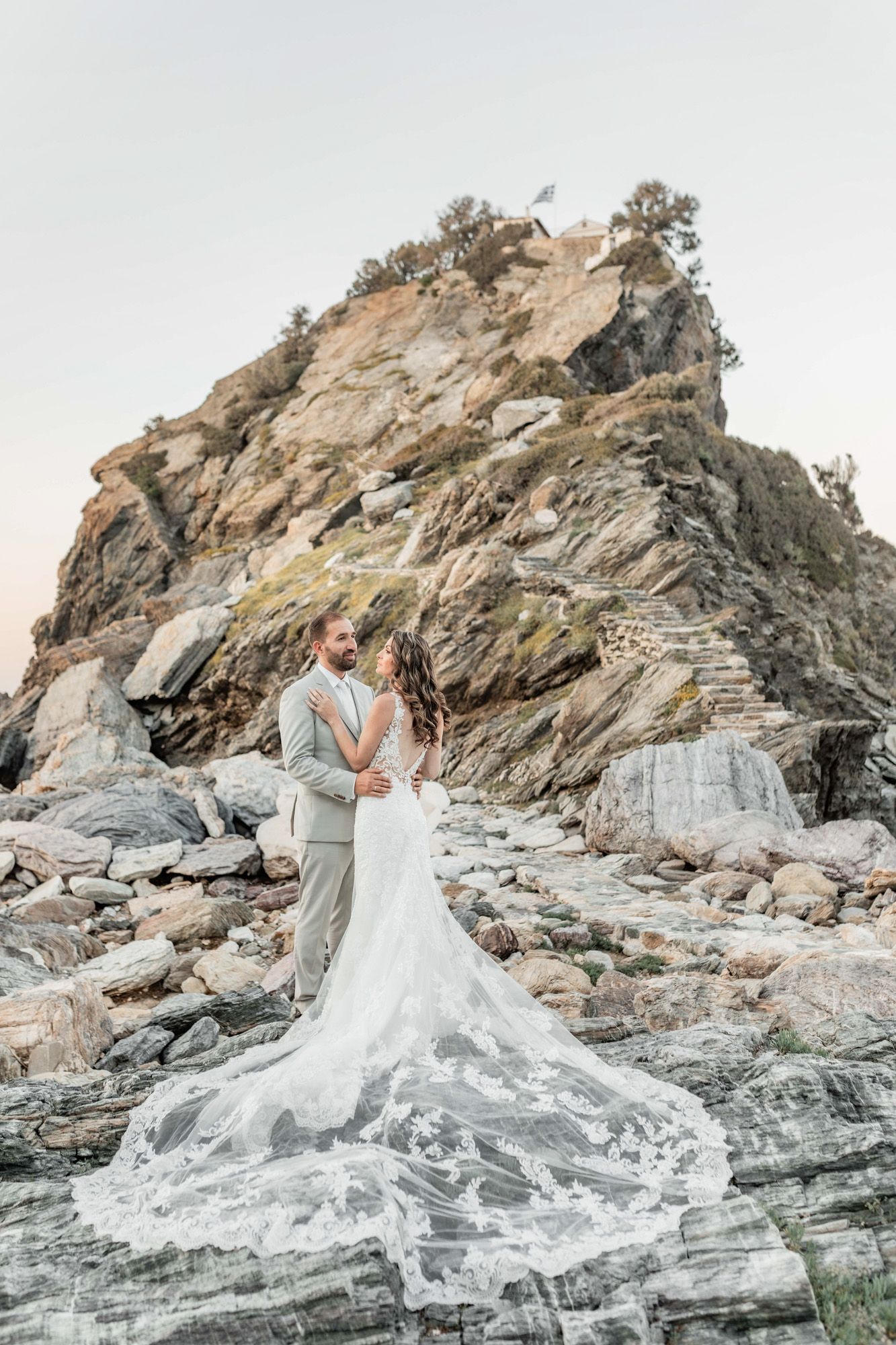 A long flowing wedding dress train drapes over coastal rocks as a couple embraces.
