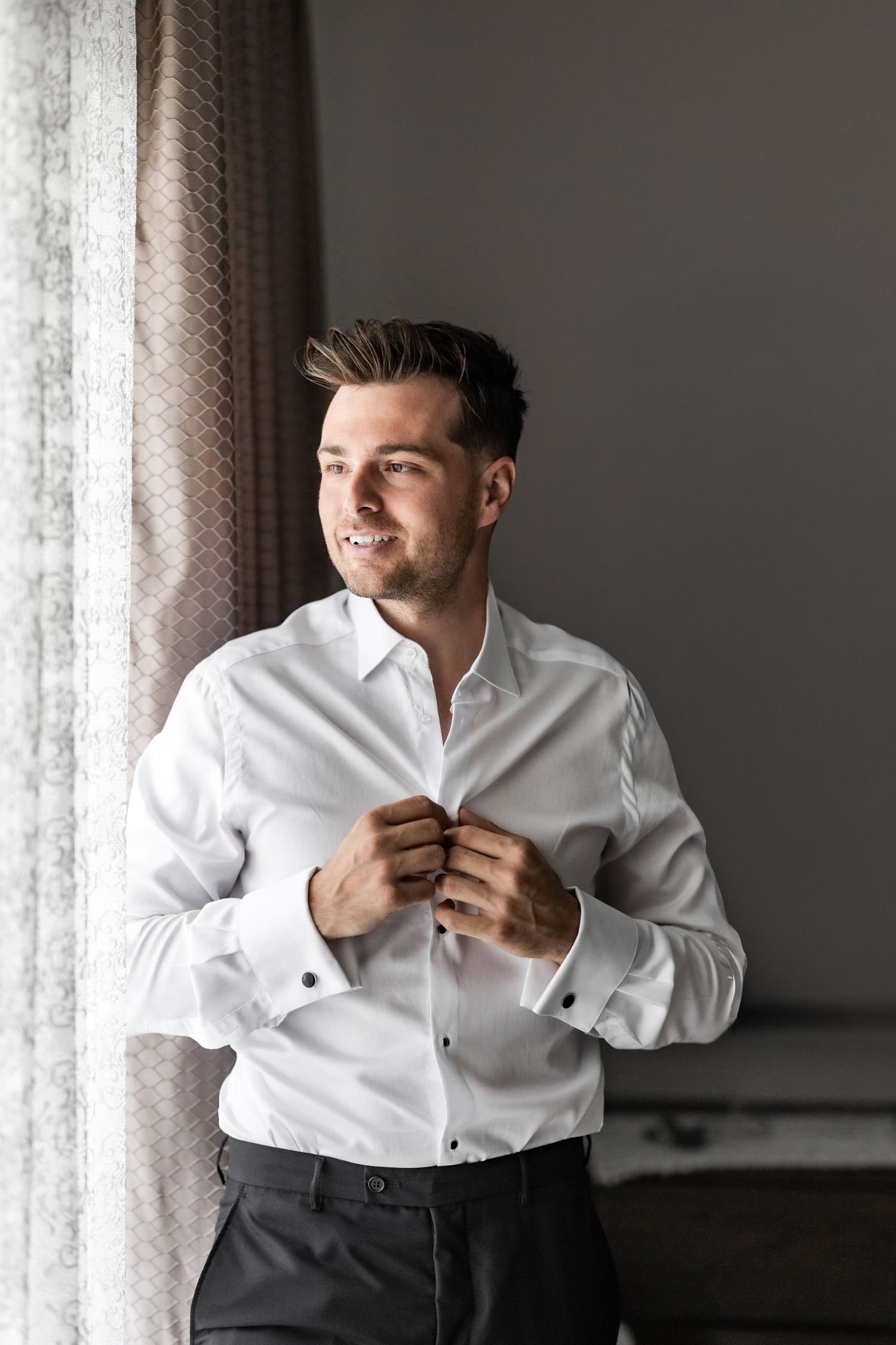 Groom in white dress shirt adjusts his cuffs while getting ready in a moody room with natural window light.