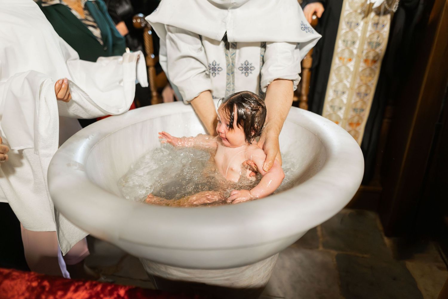 A baby is being gently lowered into a white marble baptismal font during a religious ceremony.