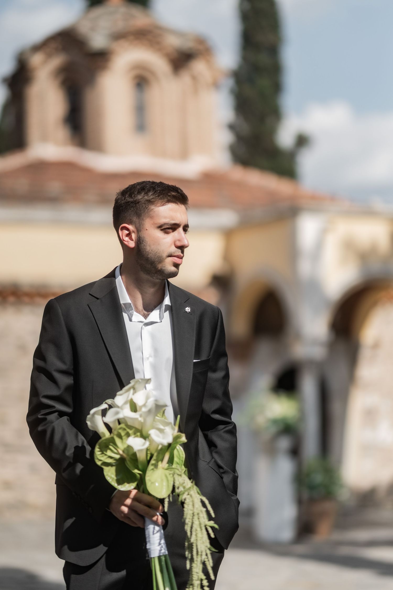 Groom in black suit holding white floral bouquet outside a historic church on his wedding day.