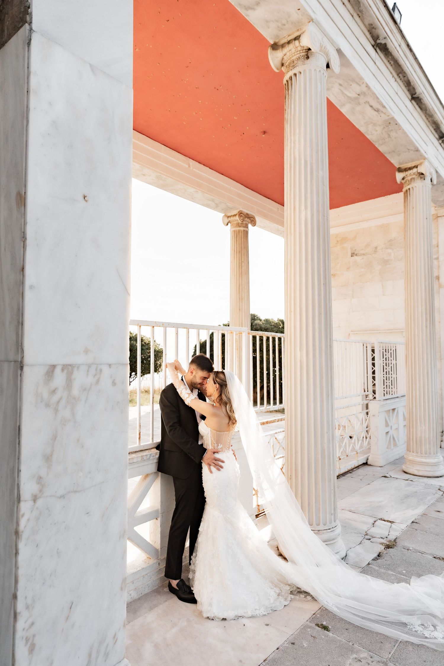 Bride and groom share romantic moment between white marble columns at elegant neoclassical venue.