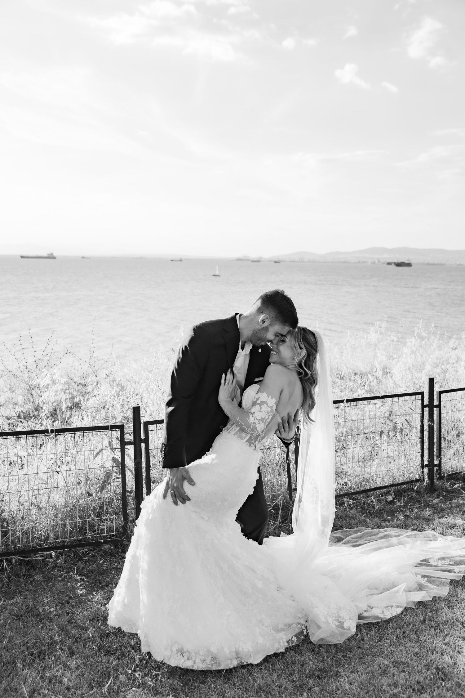 Black and white photo of a bride and groom sharing a romantic kiss by a fence with ocean views on their wedding day.