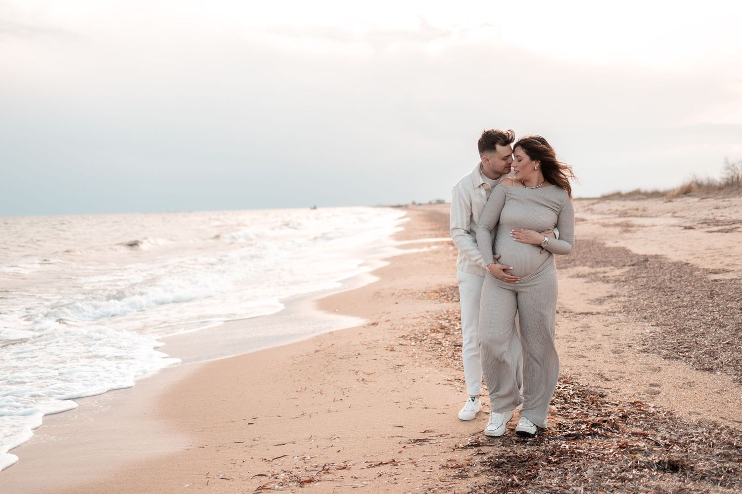 A couple walks along a serene beach shoreline at sunset with waves gently rolling onto the sand.