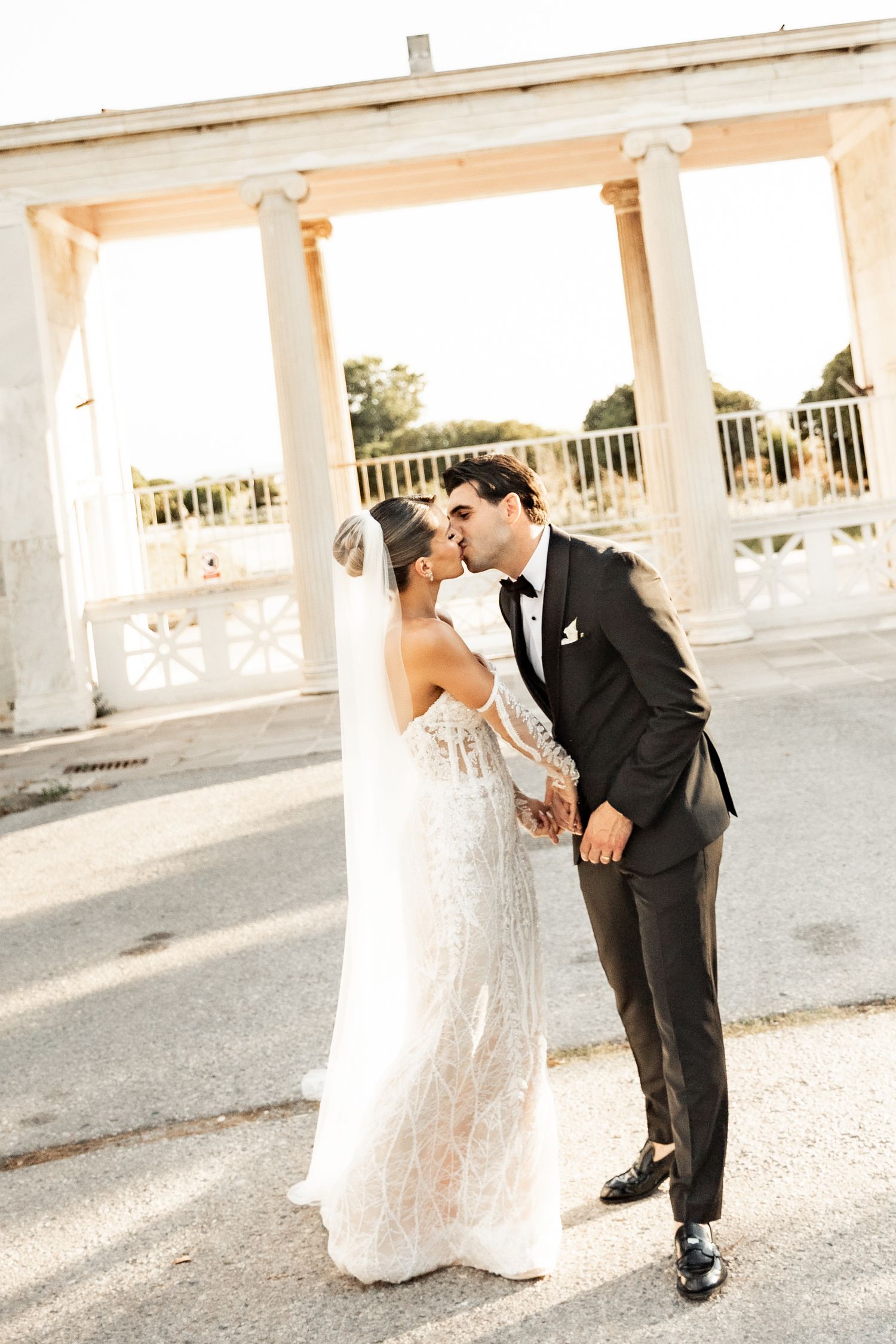 Bride and groom share a romantic kiss outdoors near elegant white columns at their wedding venue.