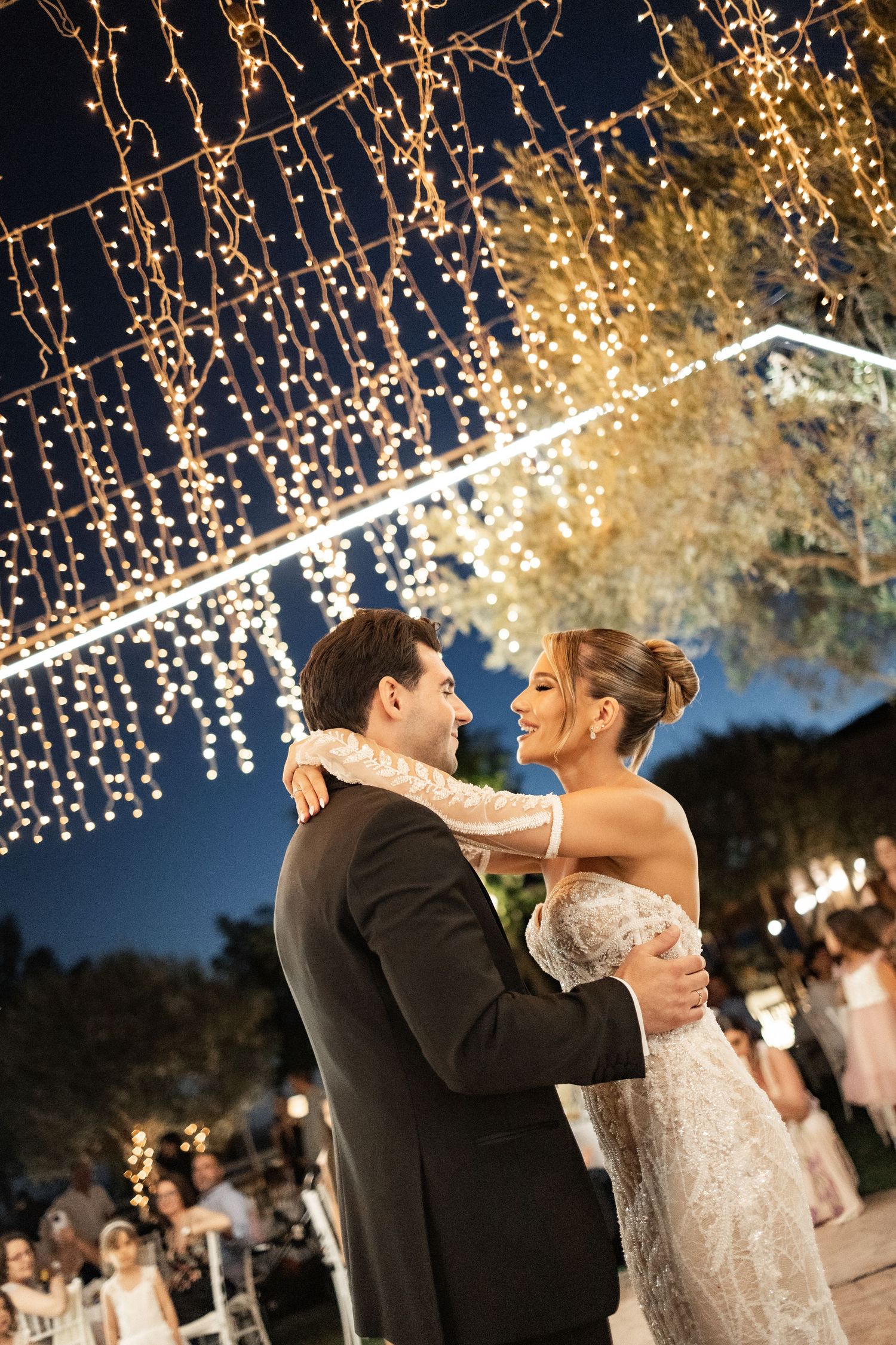 Bride and groom share first dance under twinkling fairy lights at elegant outdoor evening wedding reception.