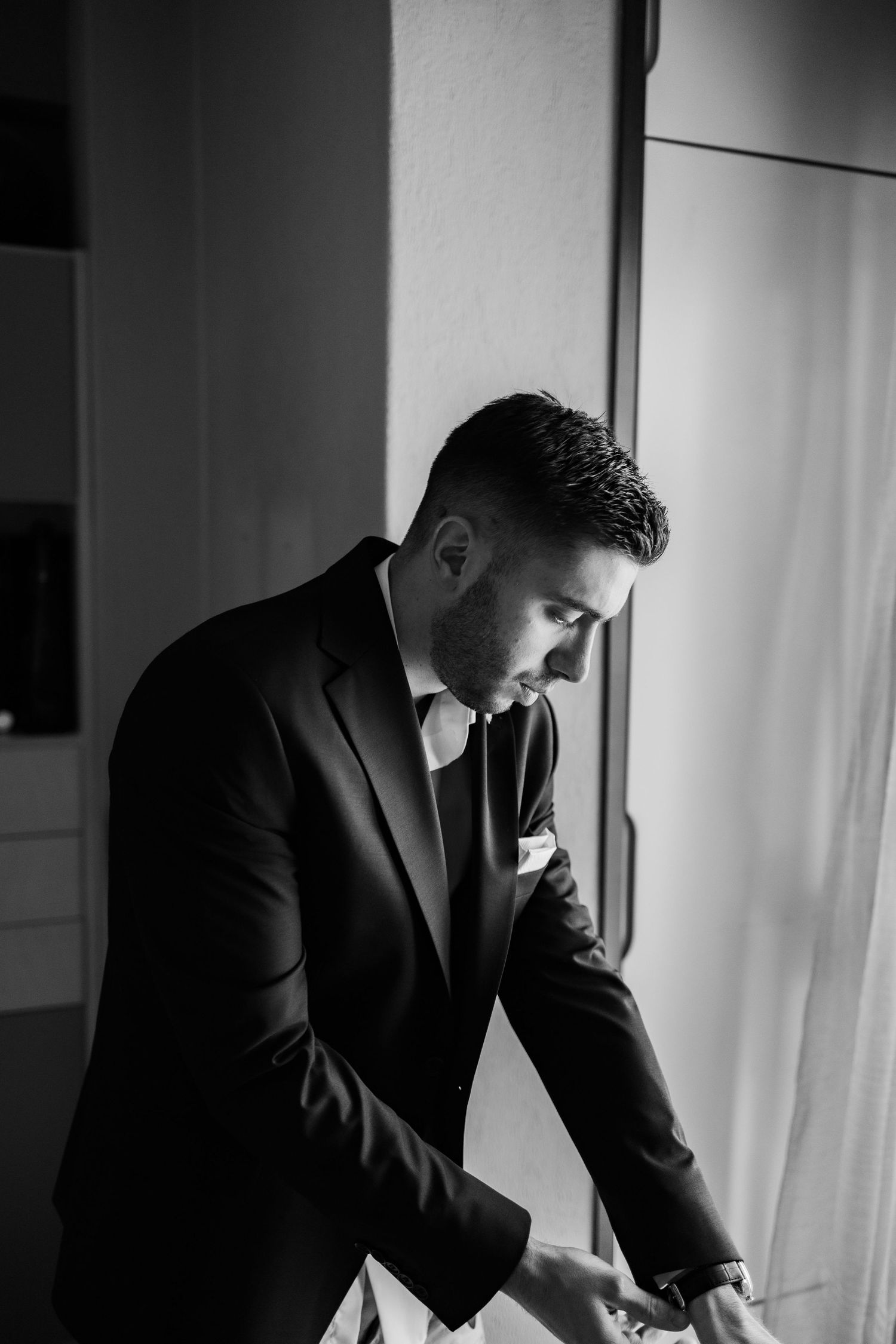 Black and white photo of a well-dressed groom in a suit adjusting his cufflinks by a window on his wedding day.