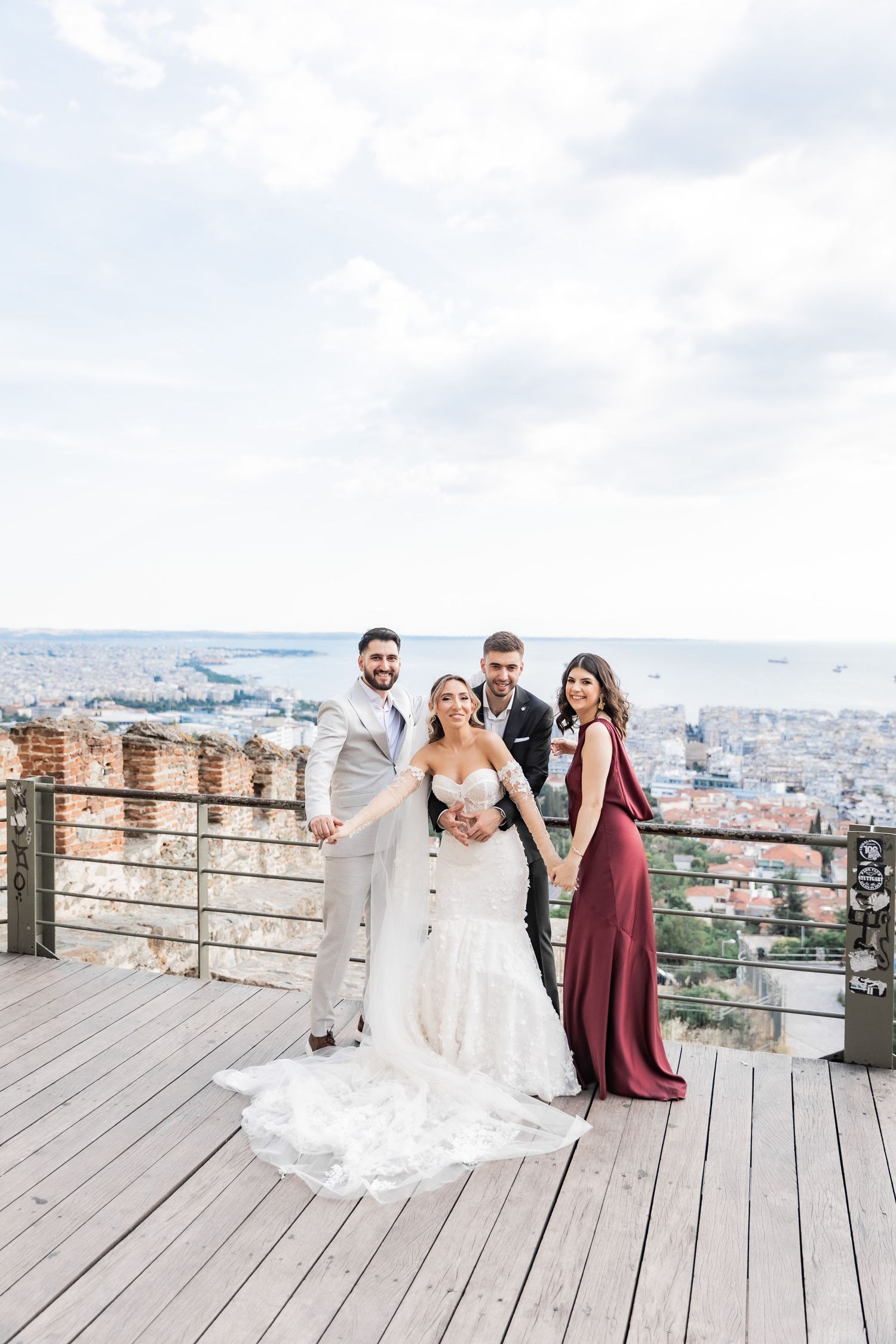 Wedding party of four poses on scenic overlook deck with city and water views behind them.