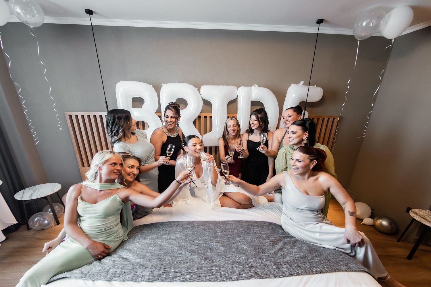 Wedding party poses playfully on bed with BRIDE letters and balloons in background.