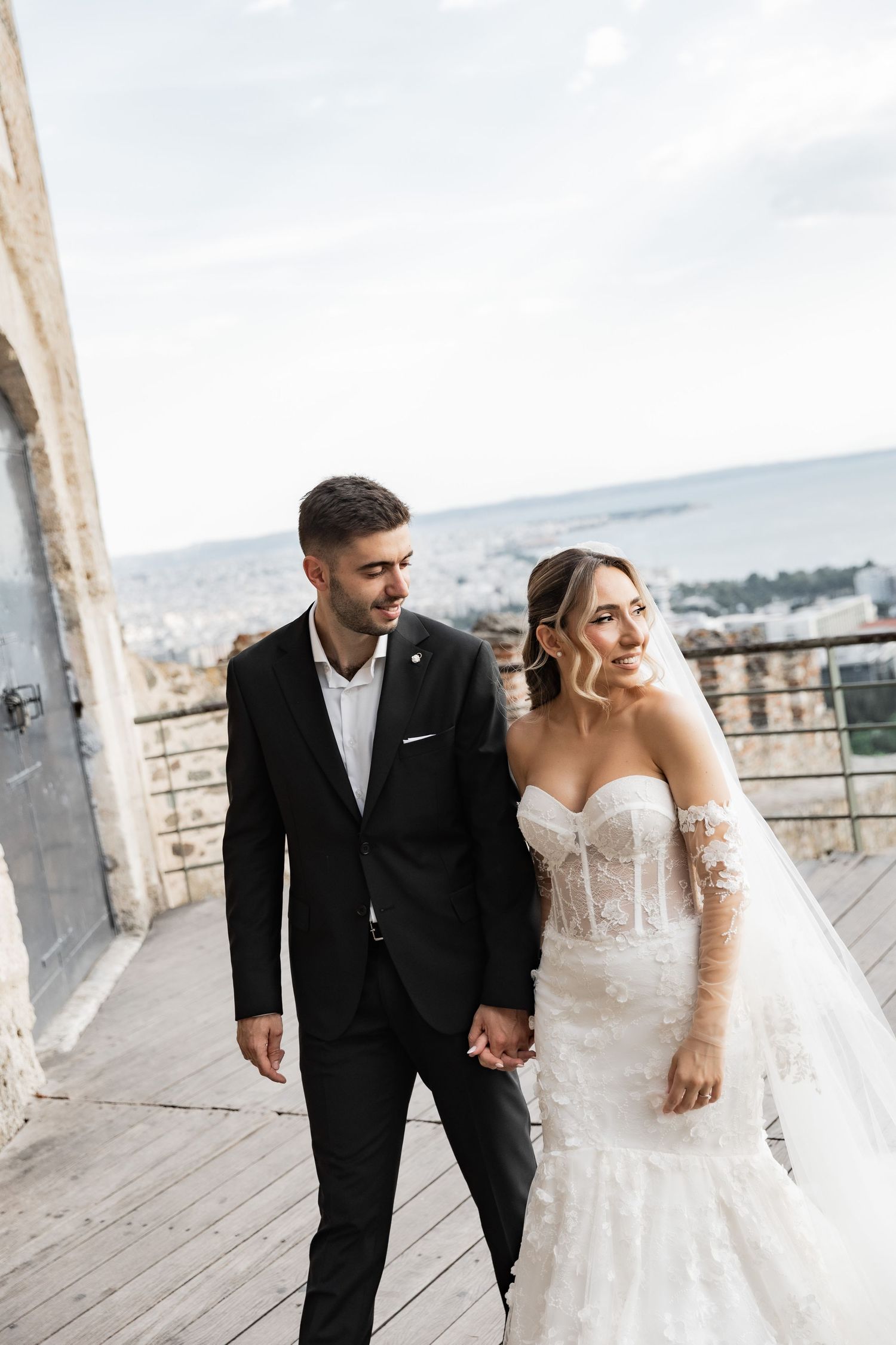 A bride in a lace gown and groom in a black suit hold hands on a scenic overlook with a city view.