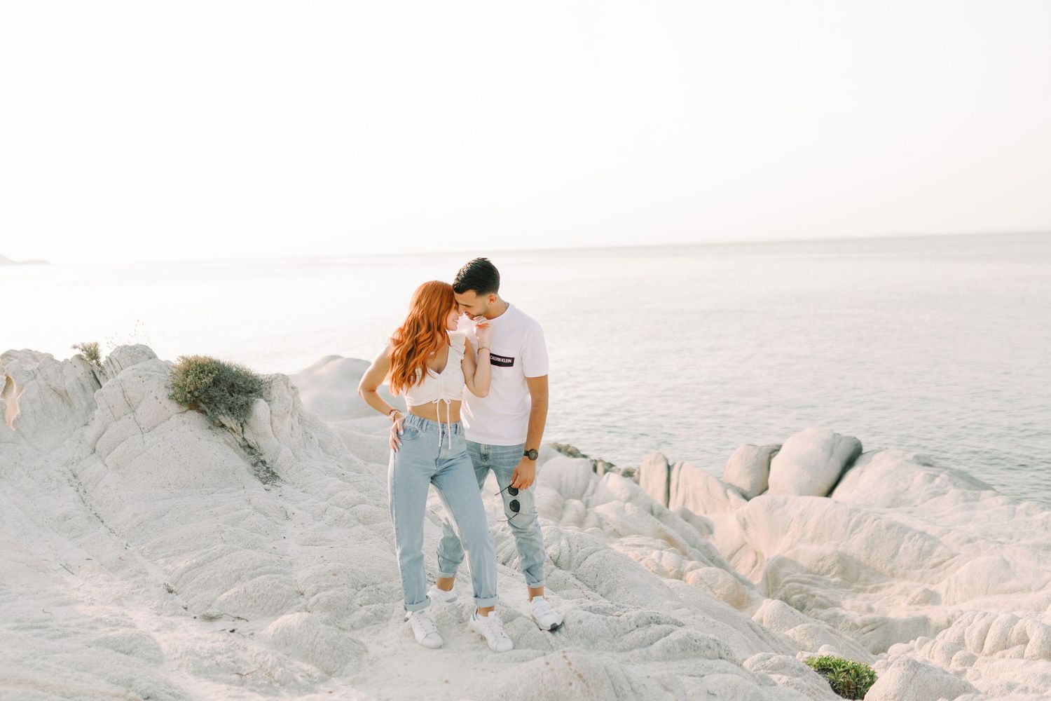 Two people walk along a scenic rocky coastline at sunset with soft waves crashing against the shore.