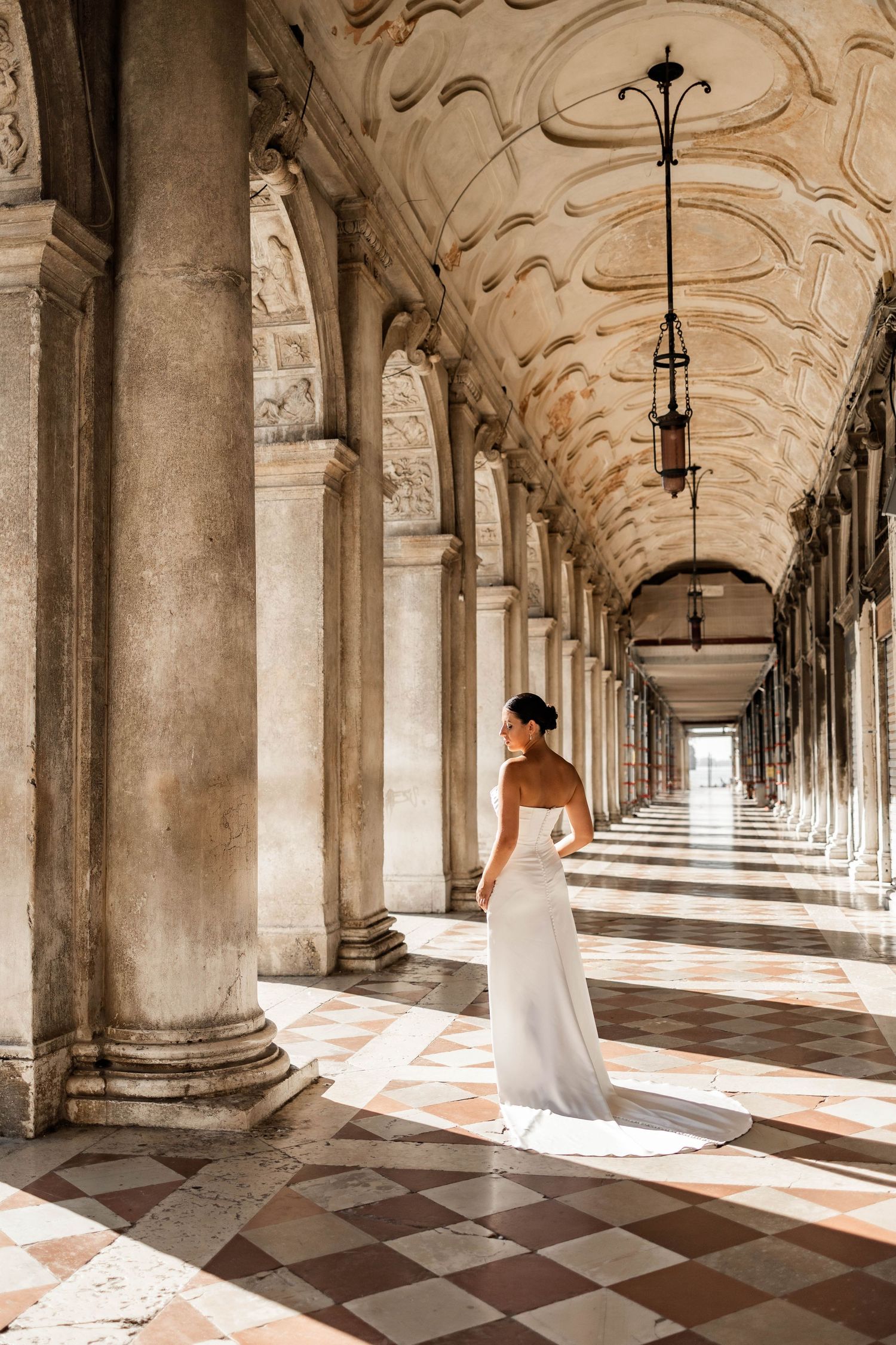 A person in a white gown walking through an ornate colonnade with checkered marble floors in Venice.