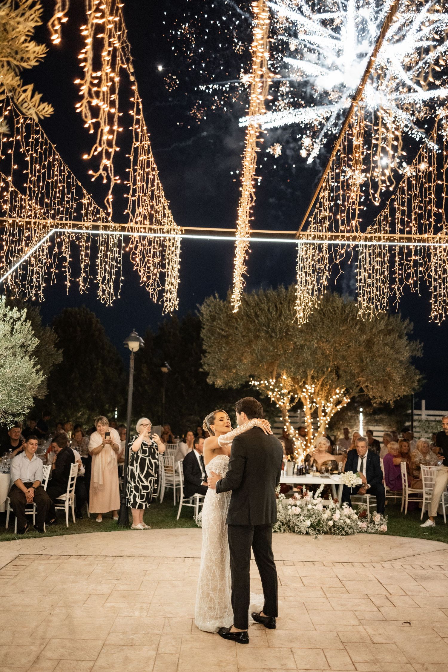Bride and groom share first dance under stunning fairy lights and fireworks at elegant outdoor wedding reception.