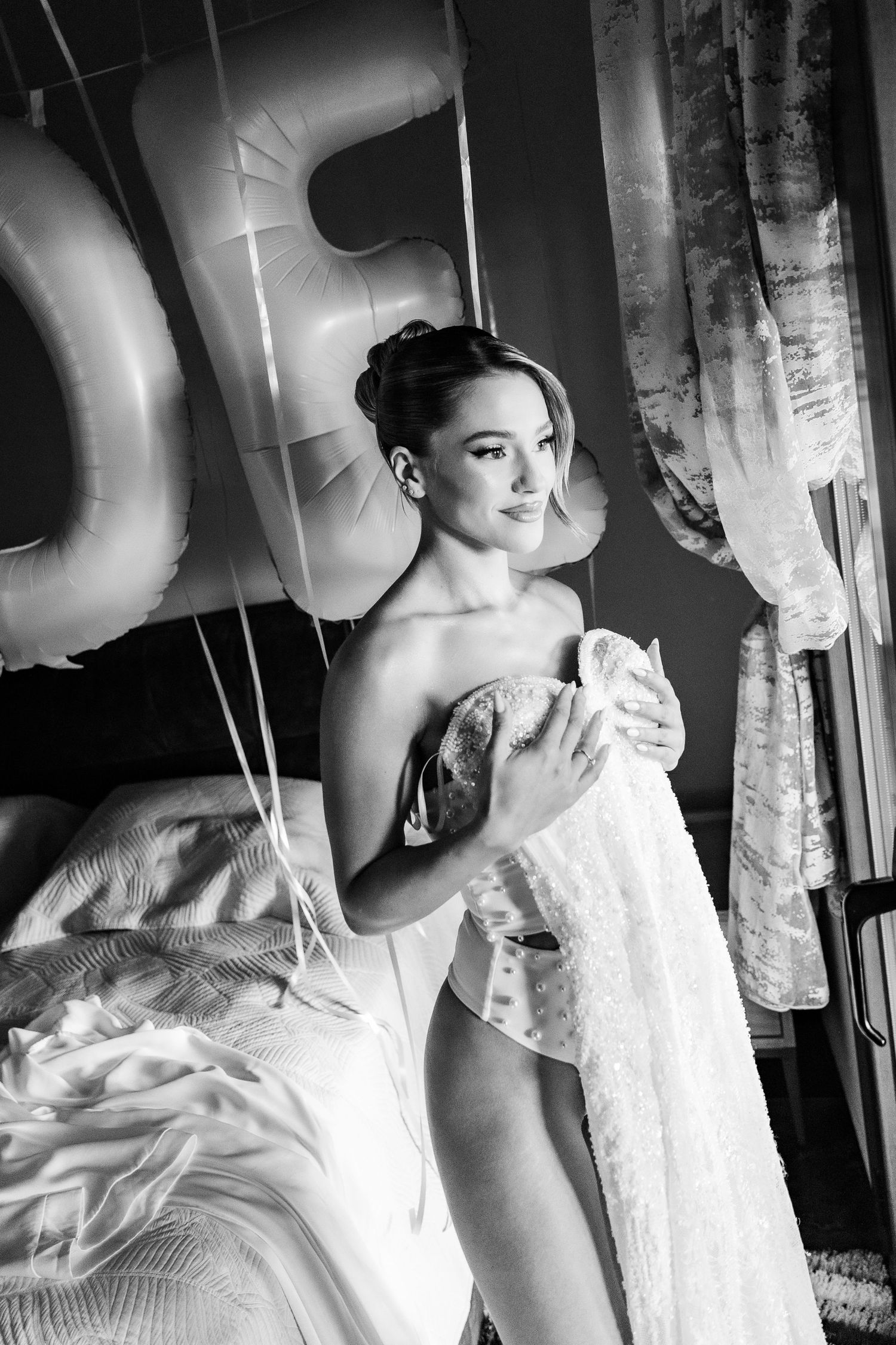 Black and white photo of a bride in a corset holding her wedding dress near balloons and curtains.