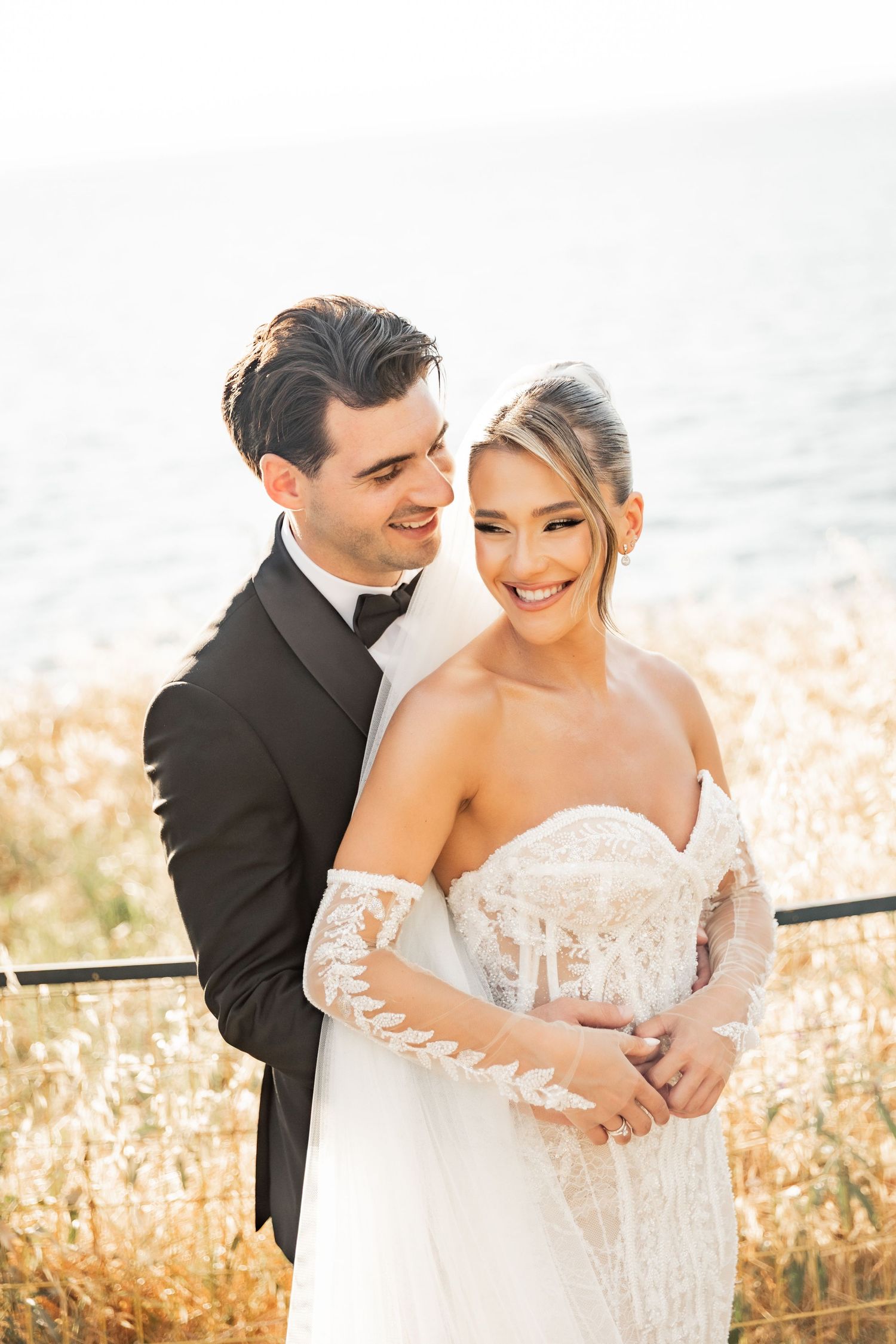 A smiling bride in lace off-shoulder gown embraced by groom in black tuxedo, posing outdoors near water at golden hour.