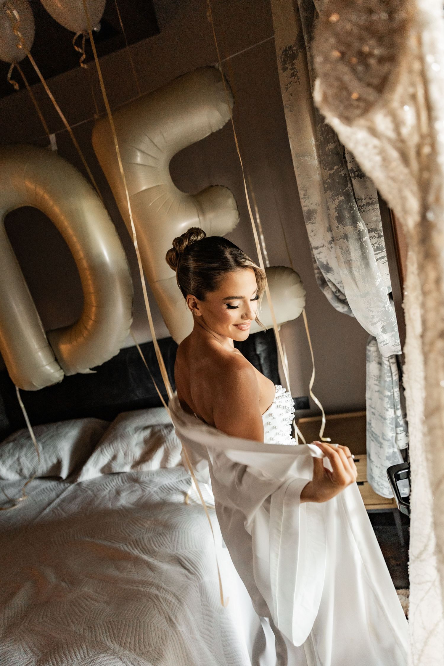 Bride in white robe sitting on bed surrounded by gold balloon letters in elegant bridal suite.