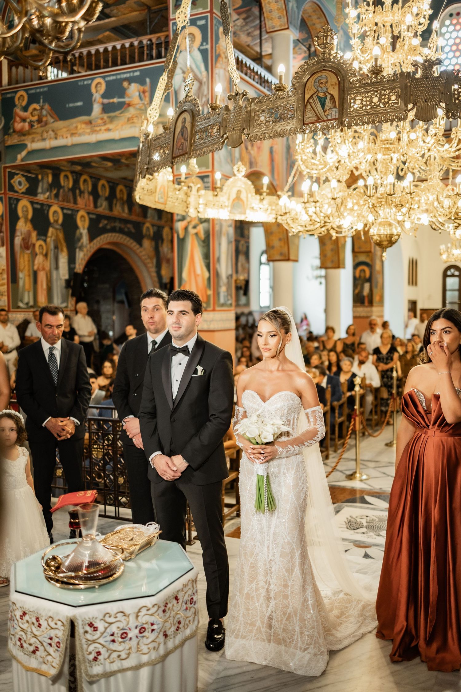 Bride and groom stand at the altar during an Orthodox church wedding ceremony with ornate chandeliers and Byzantine frescoes.