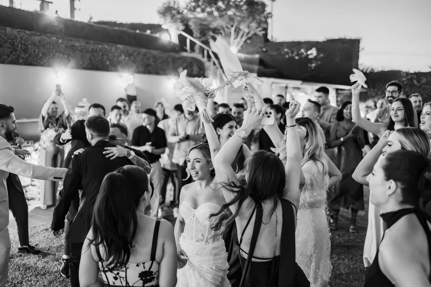 Black and white photo of a bride dancing joyfully with wedding guests outdoors at a lively evening reception celebration.