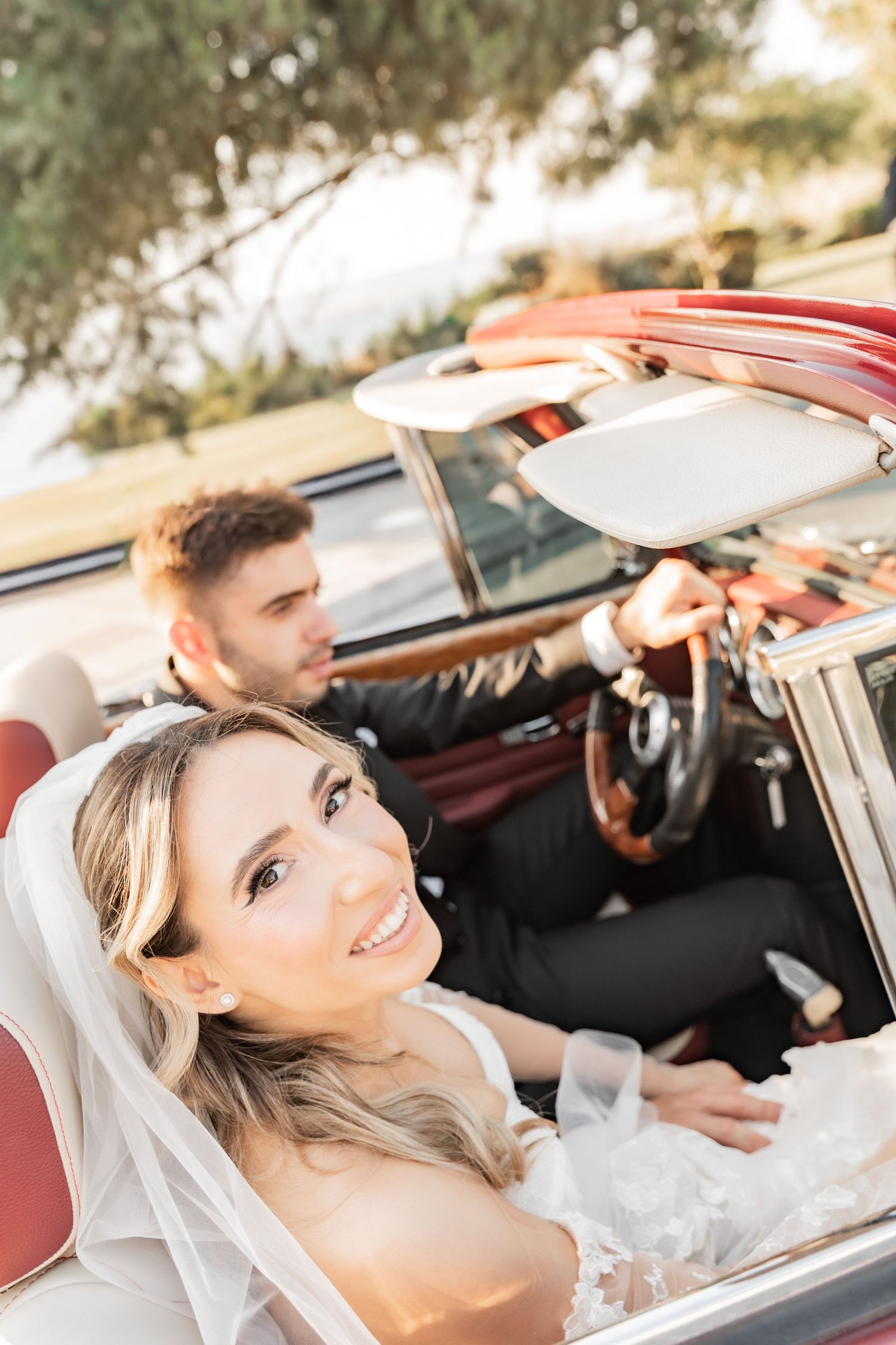 Smiling bride in white veil sits in red convertible classic car with groom in black suit driving on sunny day.