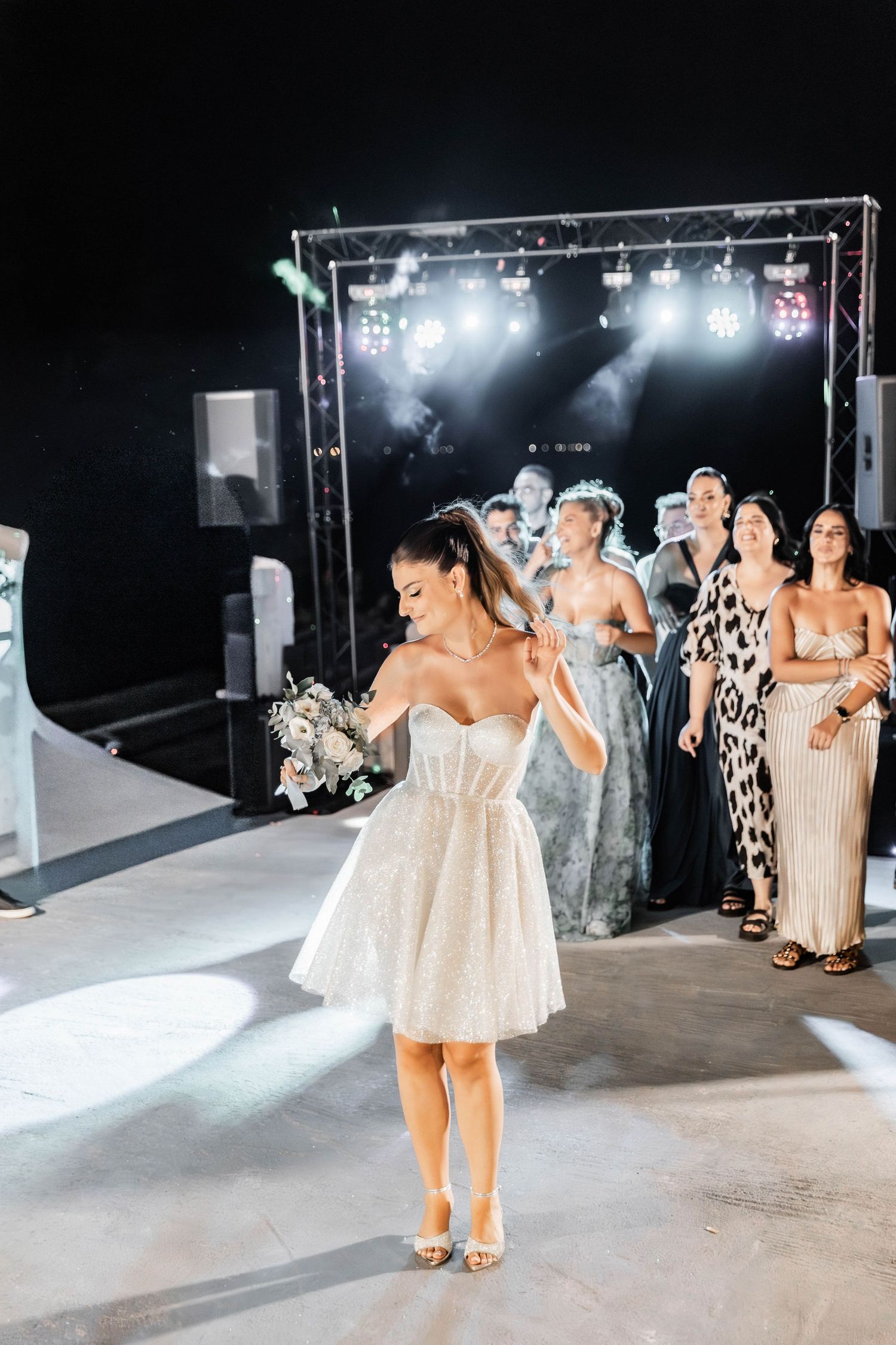 Wedding party members in matching dresses pose on a white stage during an evening celebration.