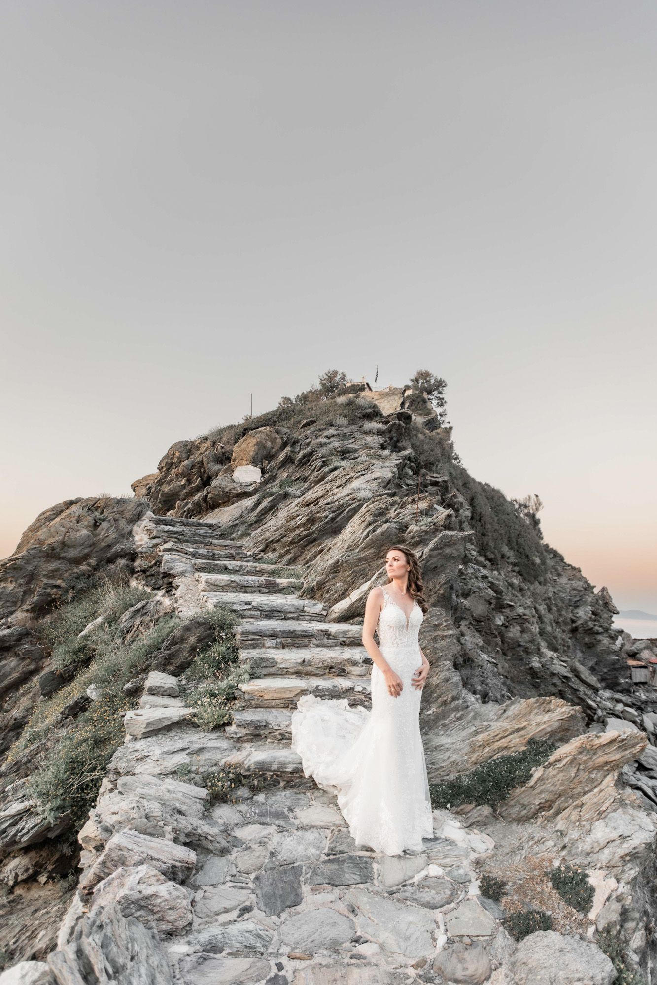 A bride in a flowing white dress stands on stone steps leading up a dramatic rocky cliff.