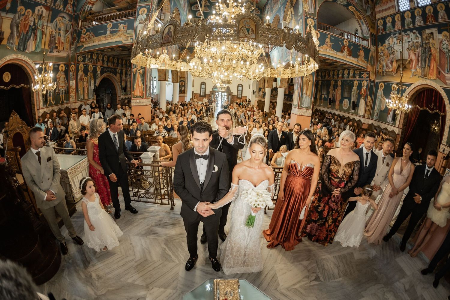 Bride and groom walk down the aisle of an ornate Orthodox church surrounded by guests during a grand wedding ceremony.
