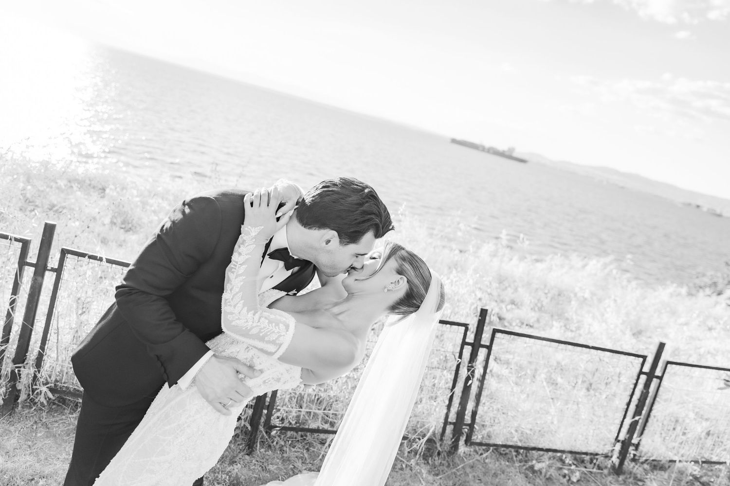 Black and white photo of a bride and groom sharing a romantic dip kiss outdoors near a waterfront fence.