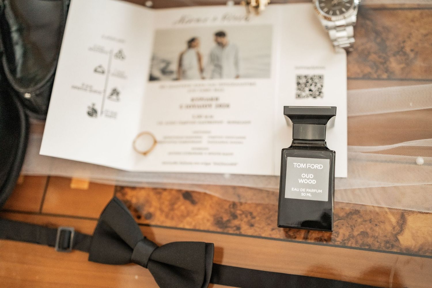 Tom Ford cologne bottle beside a black bow tie and wedding invitation on a wooden surface.