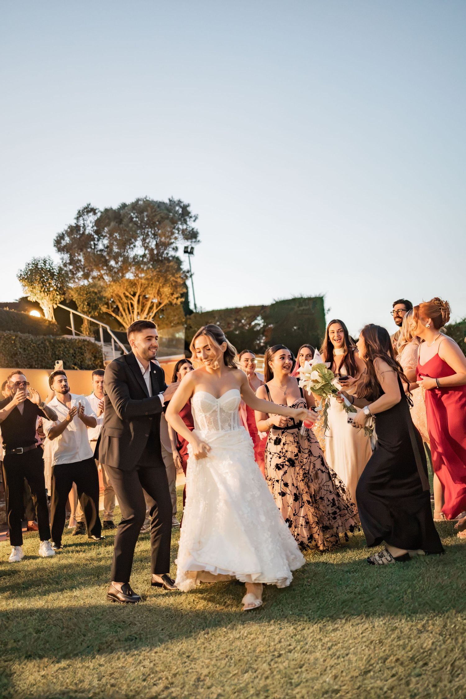 Bride and groom dancing joyfully outdoors at wedding reception surrounded by cheering guests in elegant attire at golden hour.