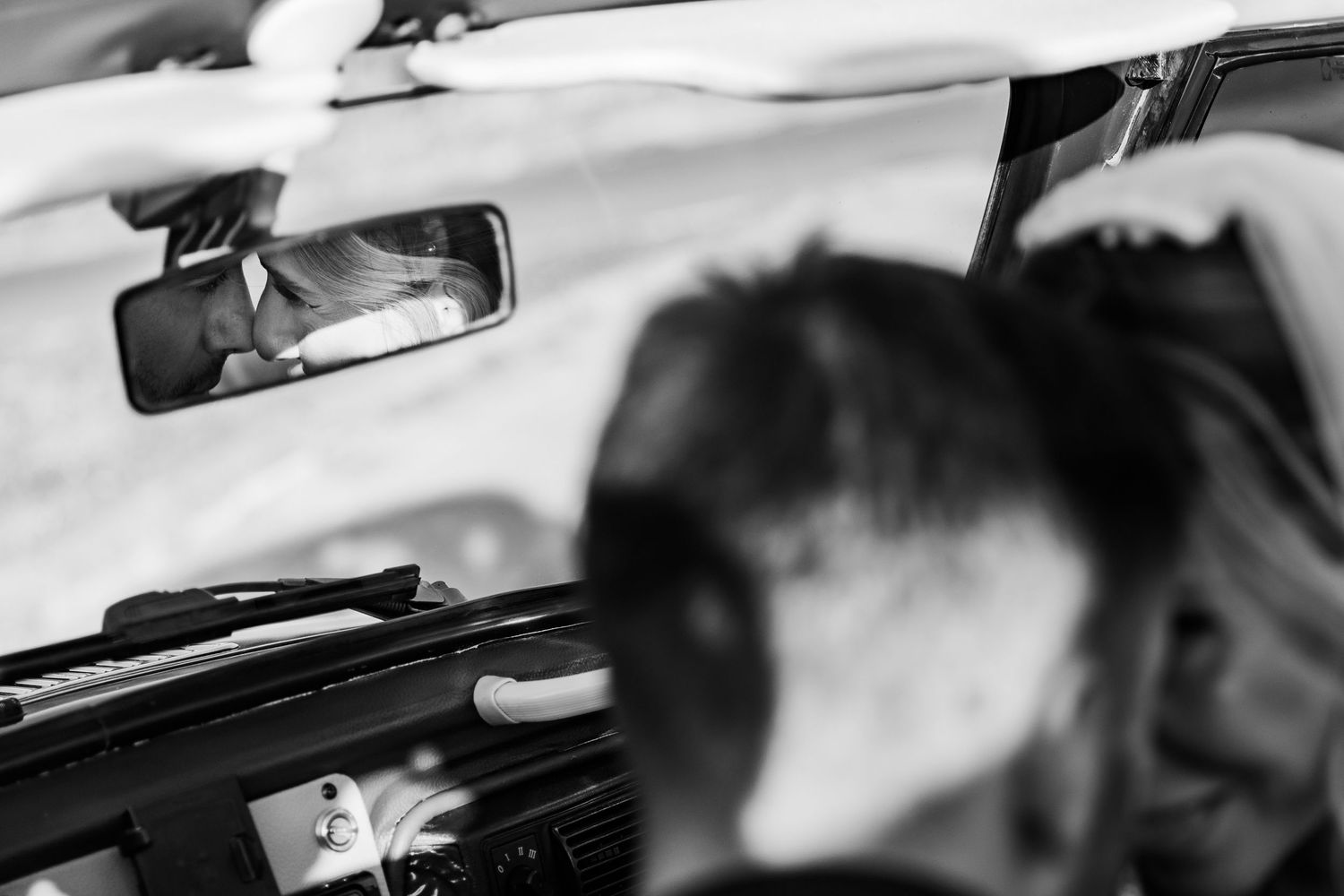 Black and white photo of a couple reflected in a car rearview mirror, capturing an intimate wedding moment.