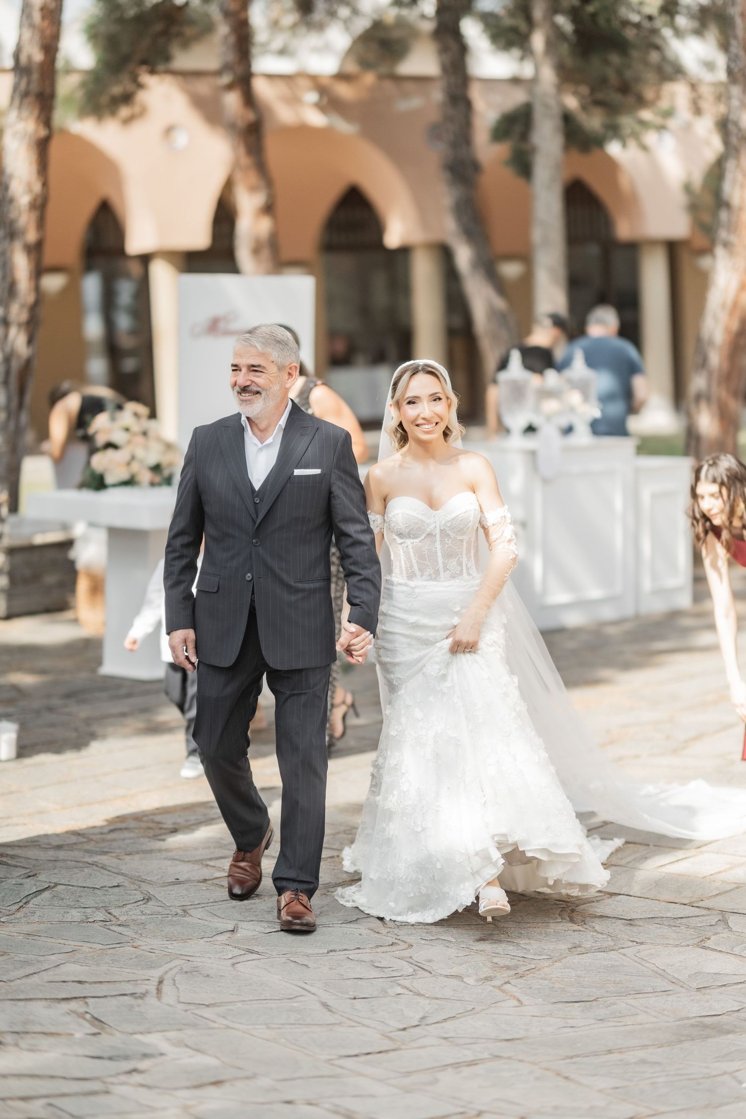 Bride in white gown walks with older man in dark suit at outdoor wedding venue.