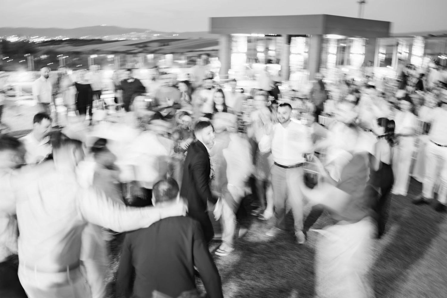 Blurred black and white photo of a large crowd dancing outdoors at a festive evening celebration.