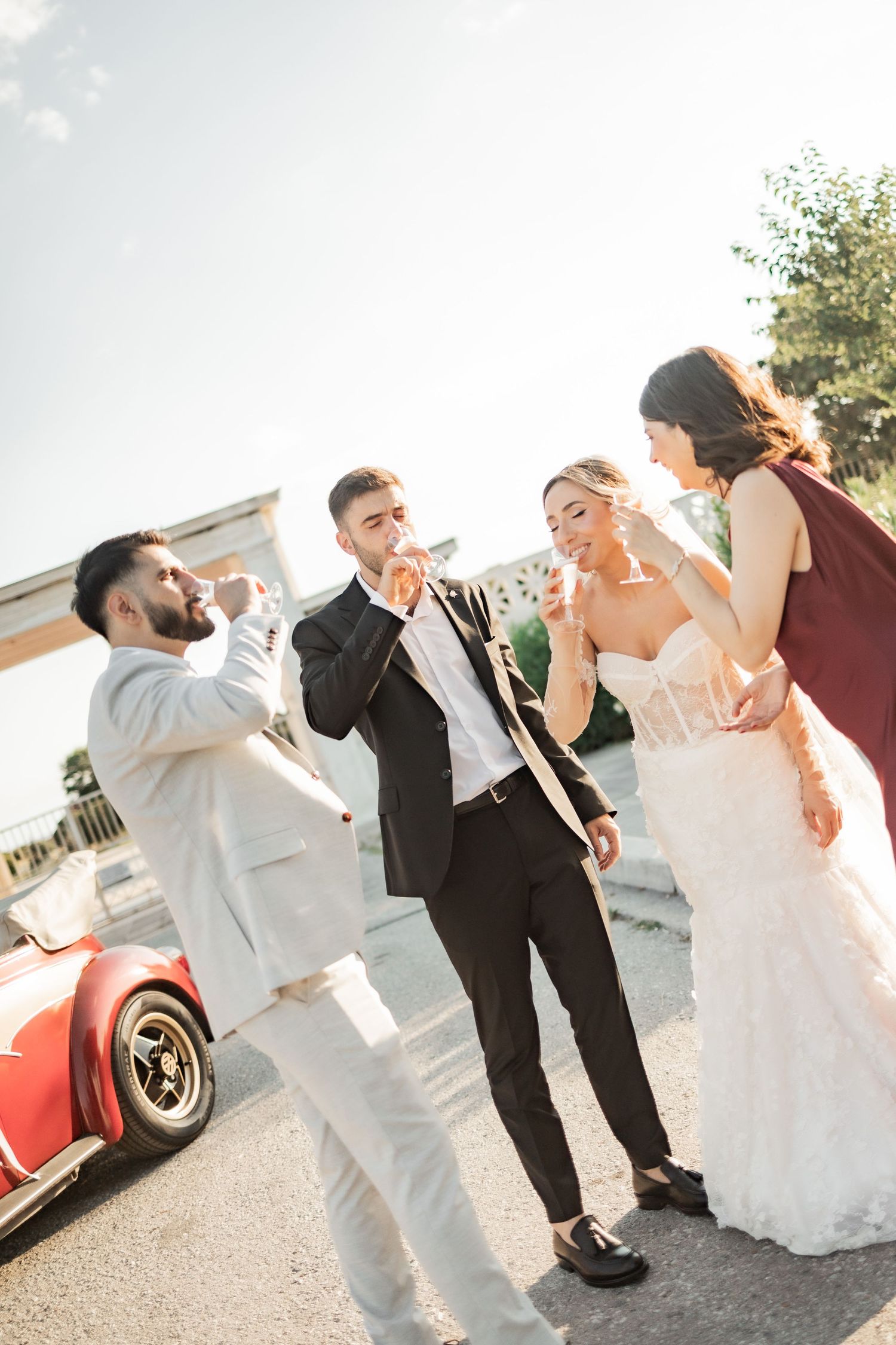 Wedding party toasting with champagne outdoors near a red vintage car on a sunny day.